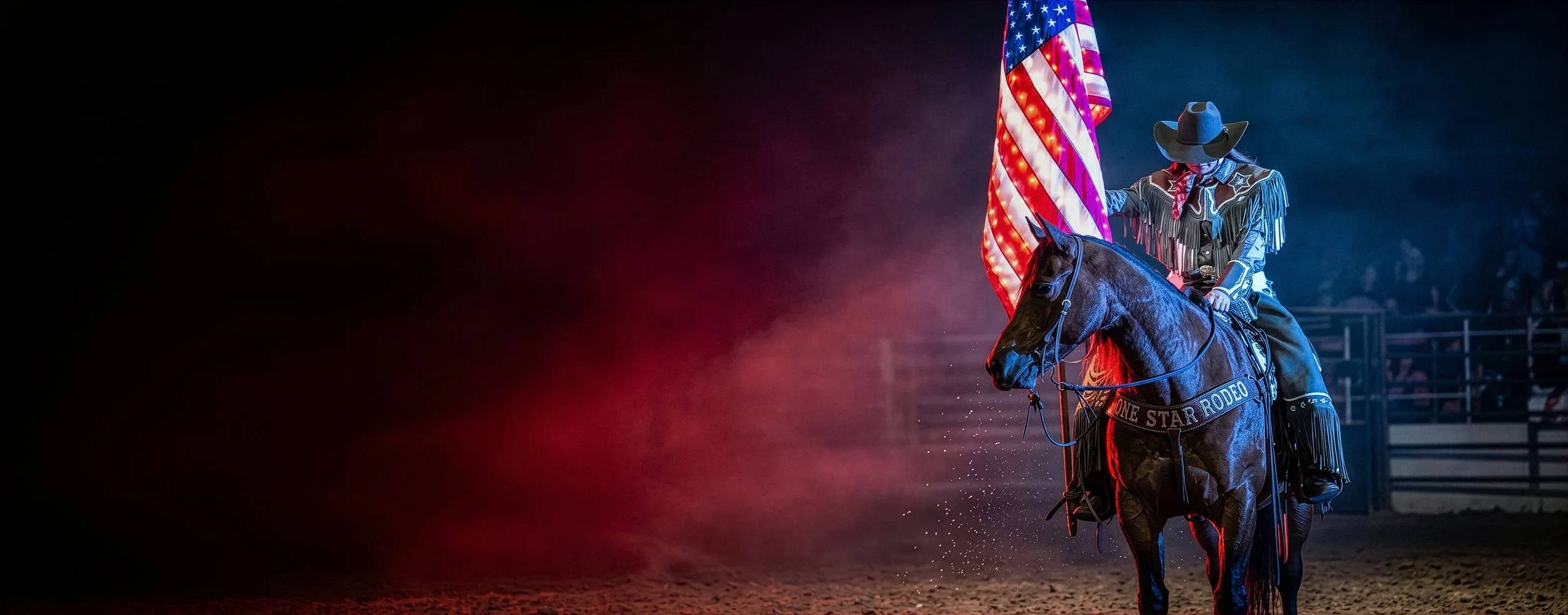 A rodeo cowboy in western attire riding a black horse, holding an American flag, in an arena with ambient lighting.