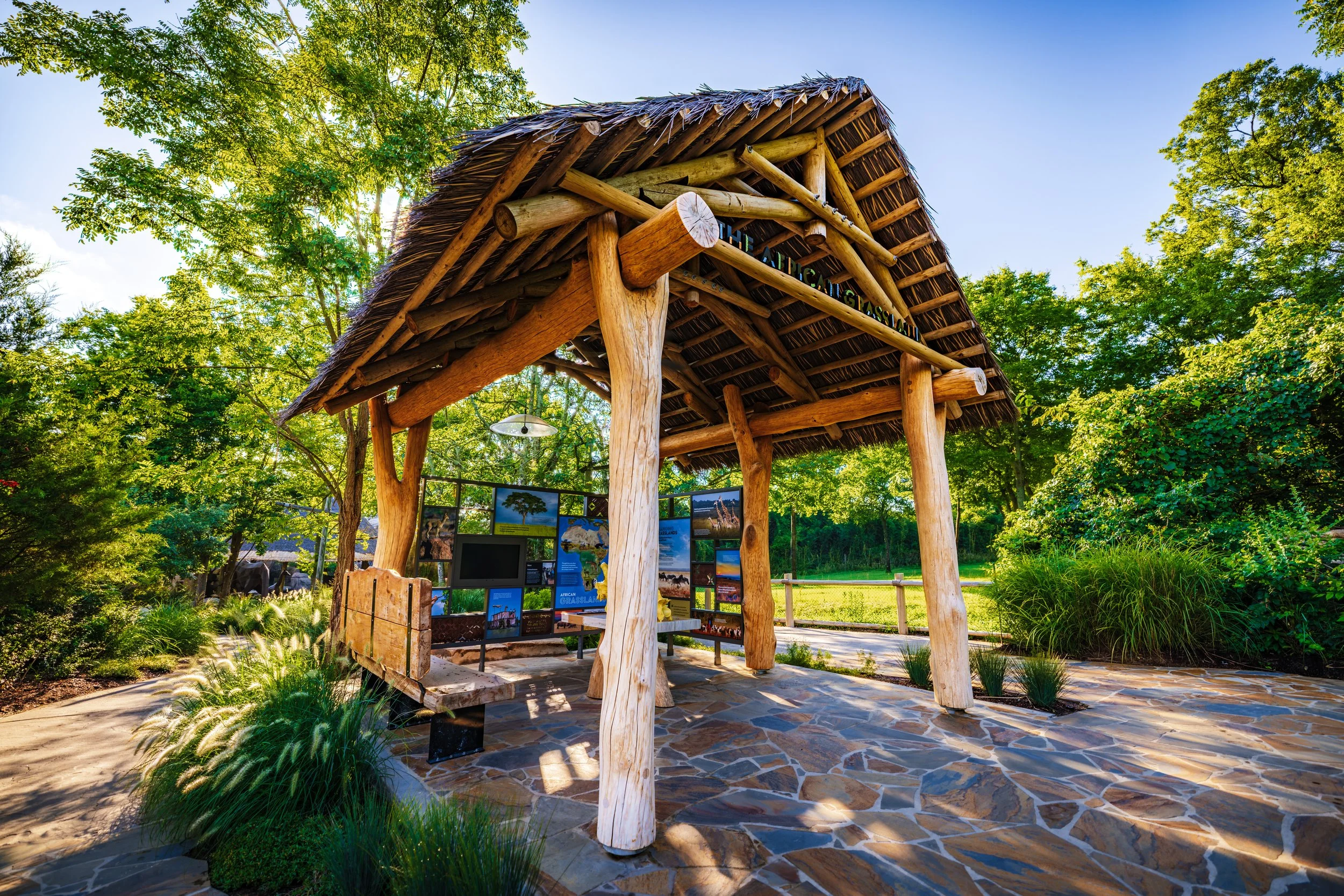 A wooden structure with a thatched roof, situated in a lush park with green trees and informational displays underneath.