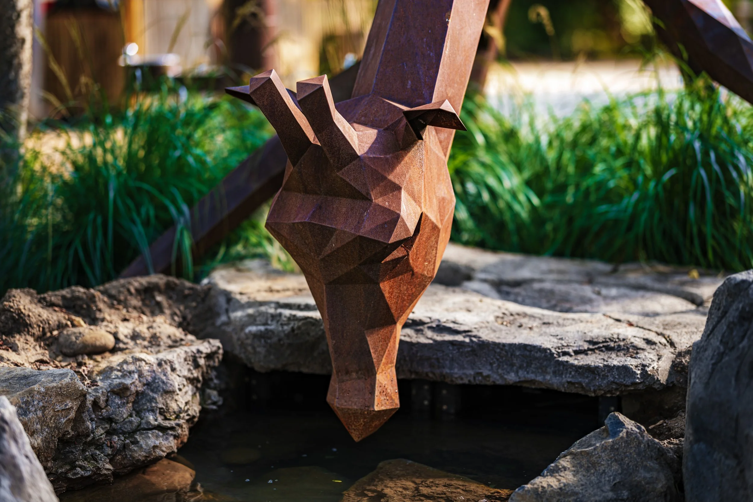 A rust-colored geometric sculpture of a giraffe's head and neck near a small water feature, surrounded by rocks and greenery in an outdoor garden setting.