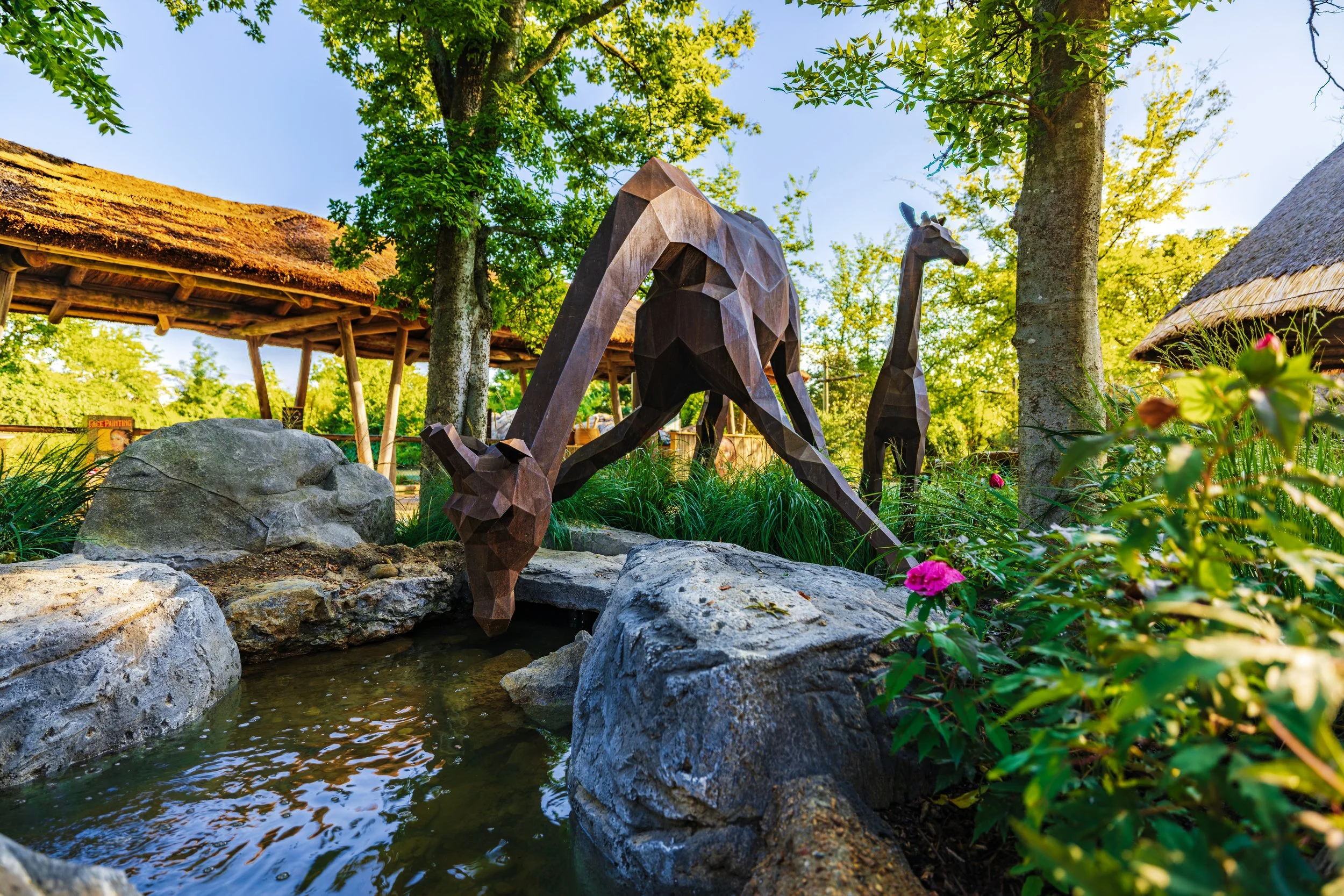 A garden scene with a metal sculpture of two giraffes, surrounded by rocks, greenery, and a small pond reflecting the sky, with a thatched-roof building in the background.