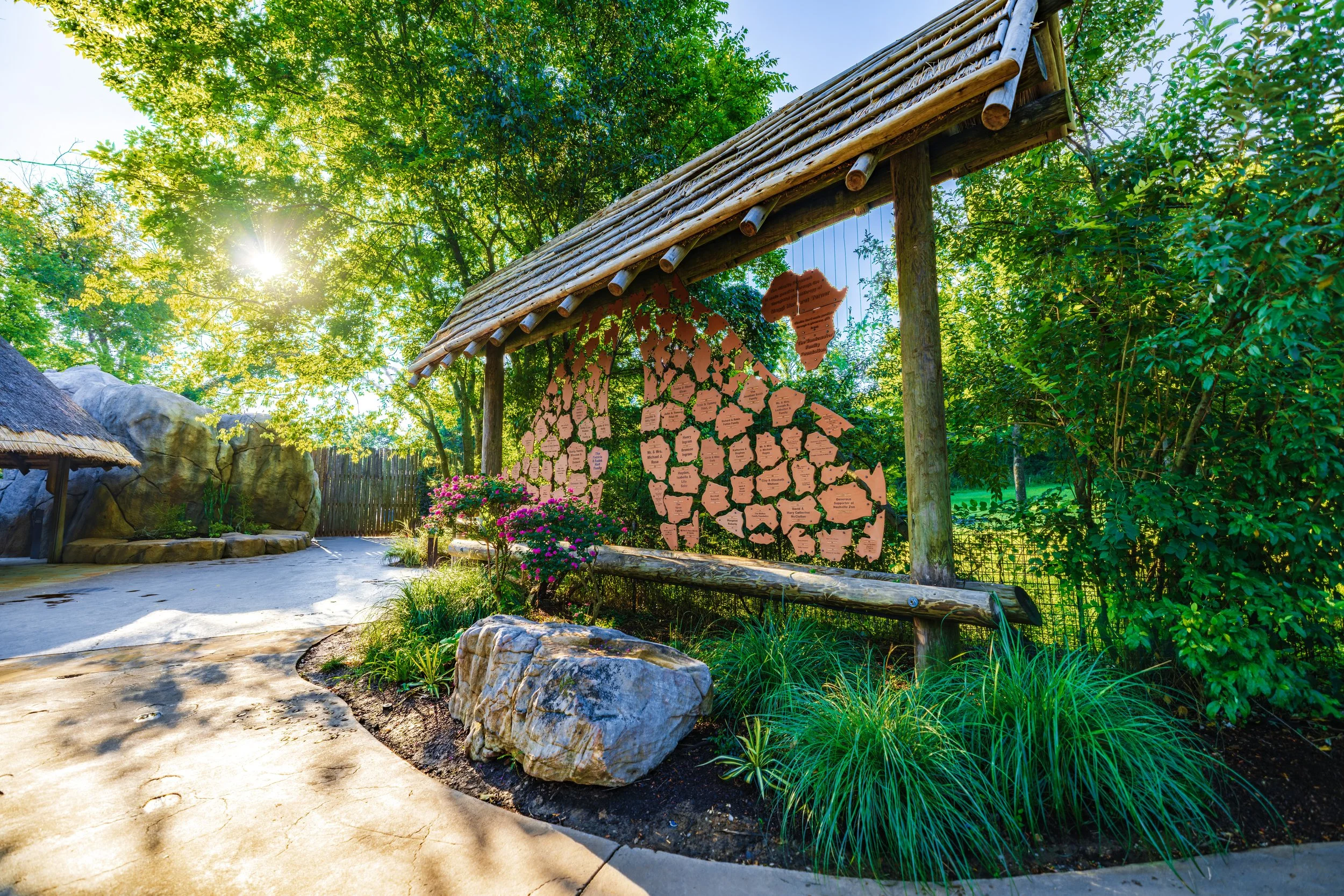 A pathway in a lush outdoor setting with a wooden signboard displaying numerous name tags, surrounded by green trees and foliage, with sunlight filtering through.