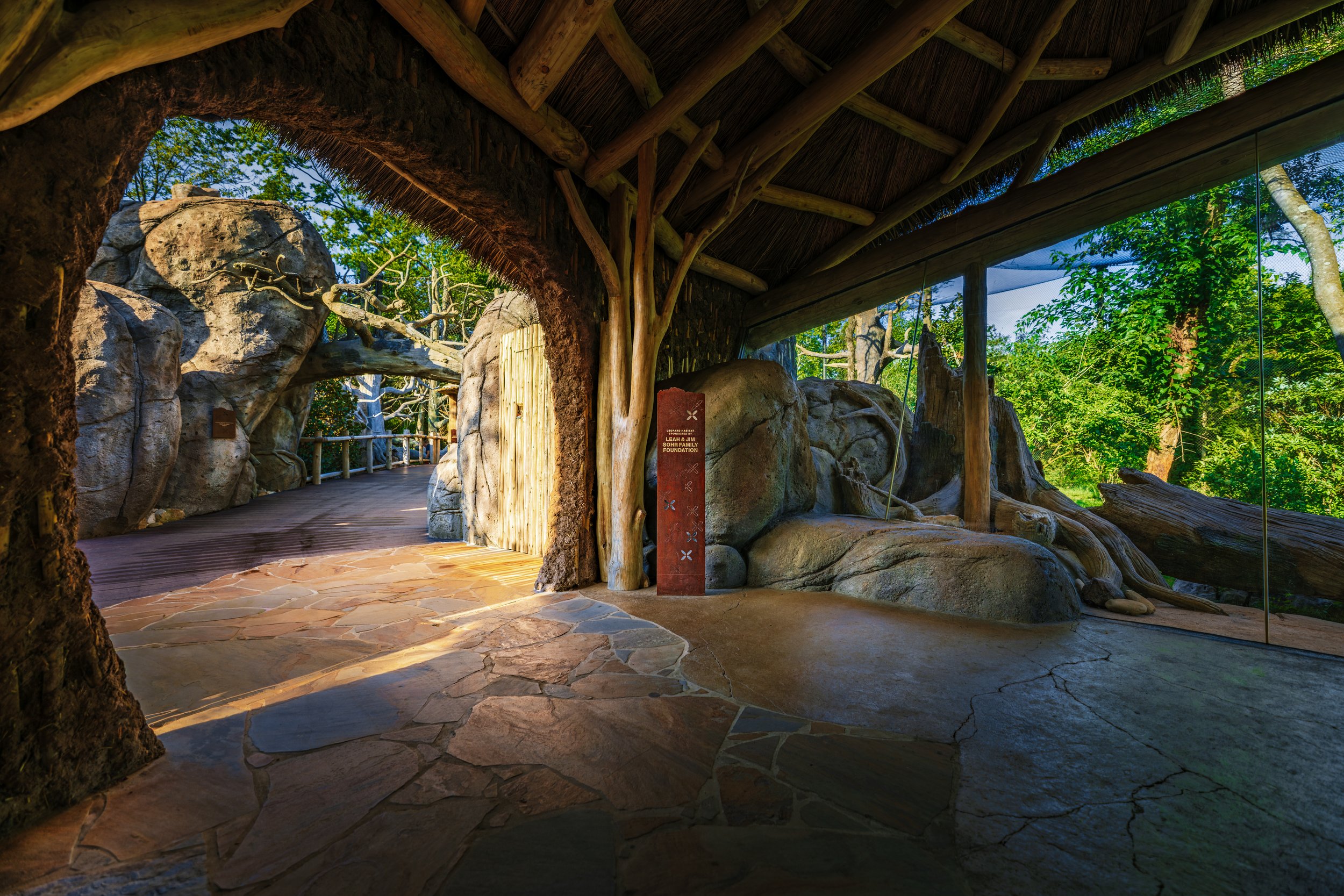 View inside an exhibit resembling a natural outdoor environment, featuring large rocks, green trees, and wooden structures, with a wooden walkway and a panel with text.
