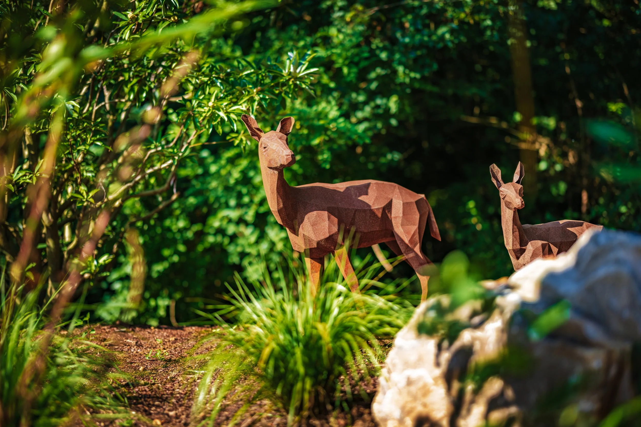Two metal animal sculptures resembling deer are placed among bushes with green leaves and grass in a garden or park setting.