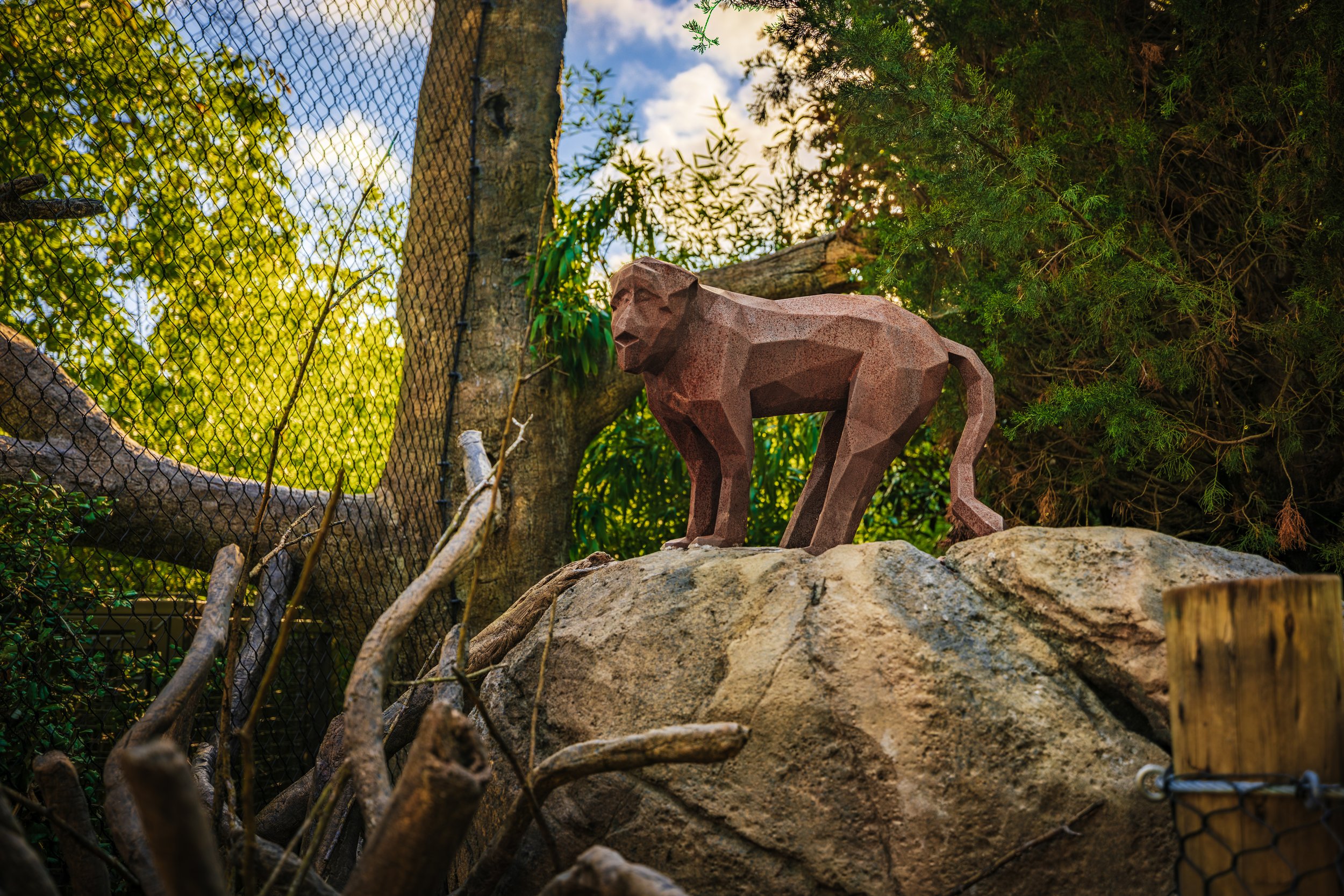 A geometric sculpture of a monkey on a large rock surrounded by trees and shrubbery, with a fence in the background and a blue sky with some clouds.