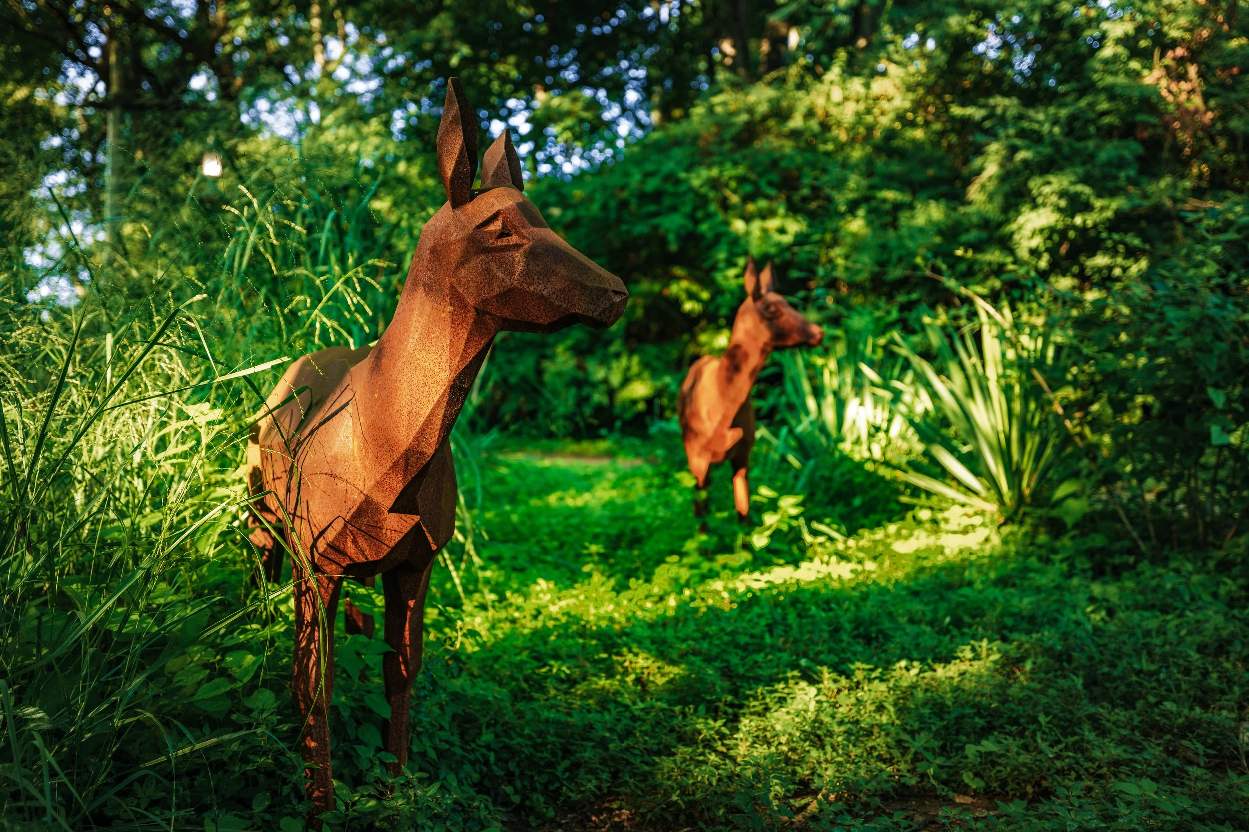 Metal animal sculptures resembling deer in a lush green park with sunlight filtering through the trees.
