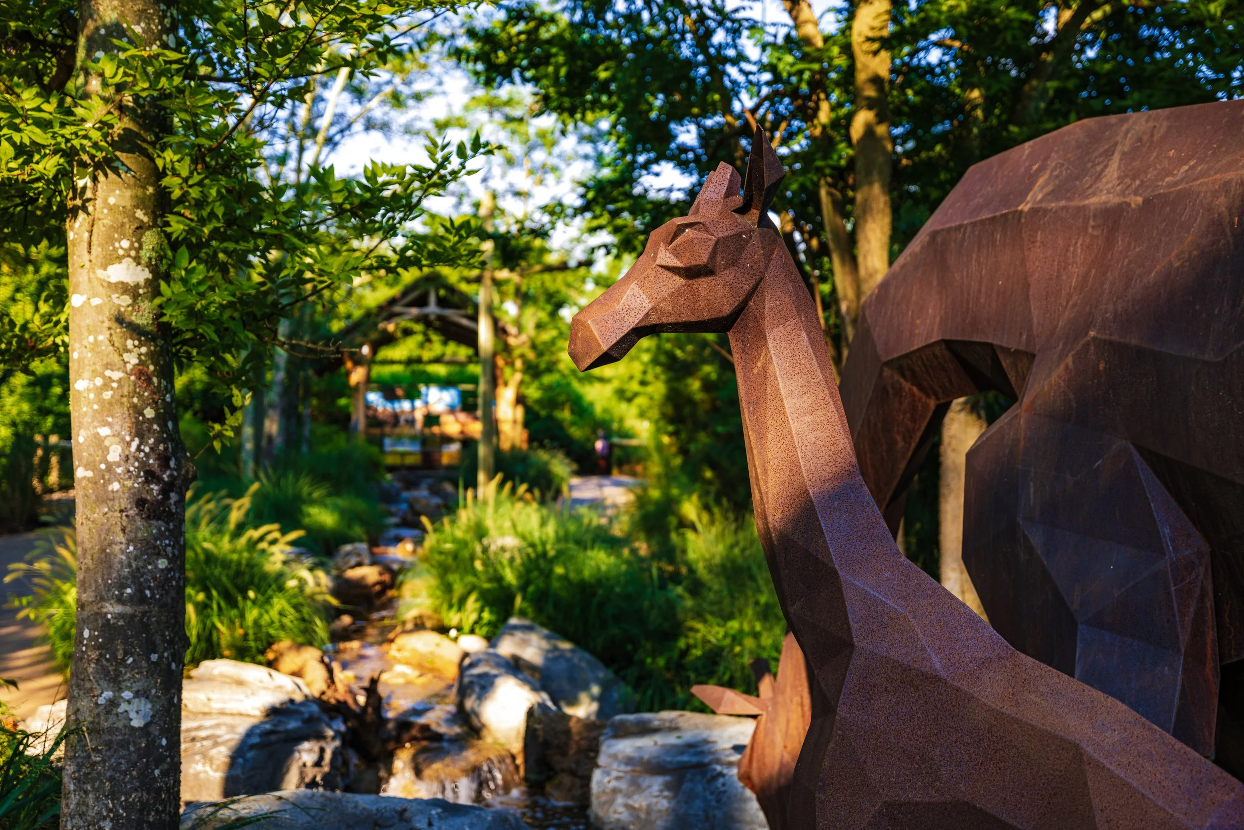 A decorative metal sculpture of a giraffe in a park with trees and a rocky stream.