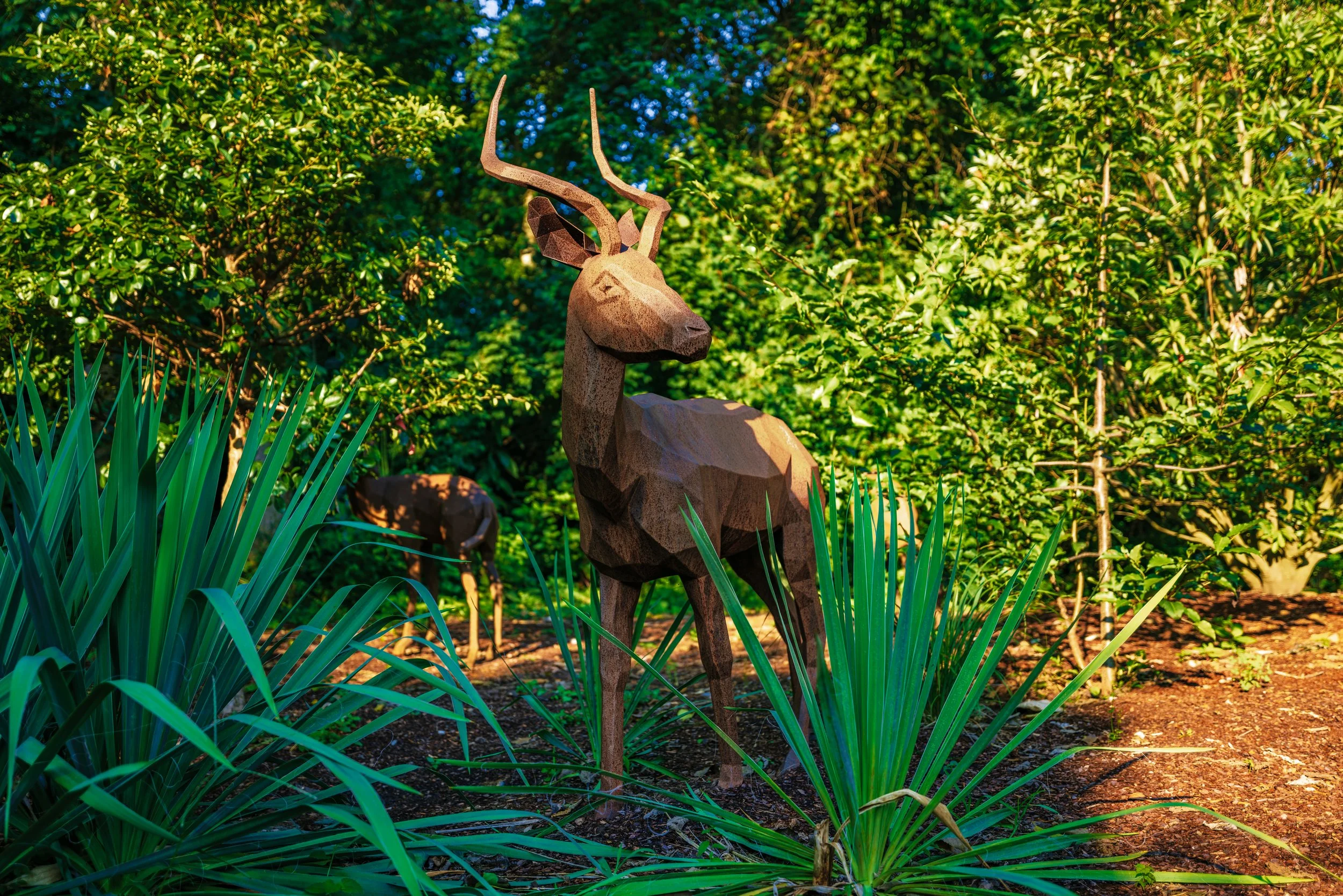 Metal sculpture of a gazelle or antelope positioned among green plants and bushes in a natural outdoor setting with sunlight.