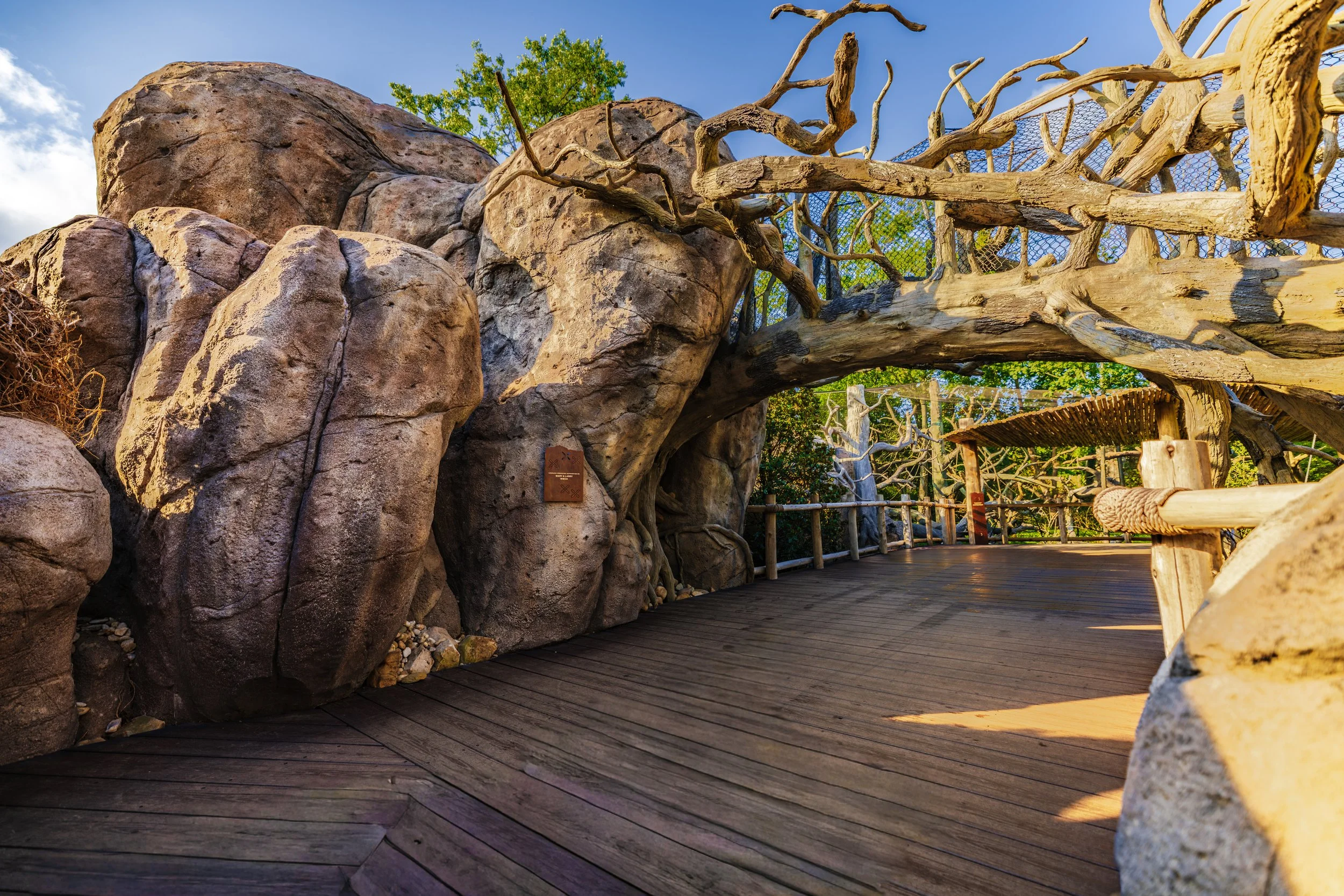 A rocky outdoor area with large boulders and leafless tree branches, a wooden walkway, and a blue sky with some clouds.