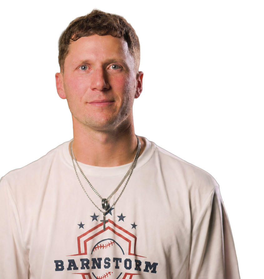 A young man with short curly brown hair, blue eyes, and fair skin, wearing a white T-shirt with a baseball-themed logo that says "Barnstorm" with a baseball graphic and stars, and layered silver necklaces with a cross pendant.