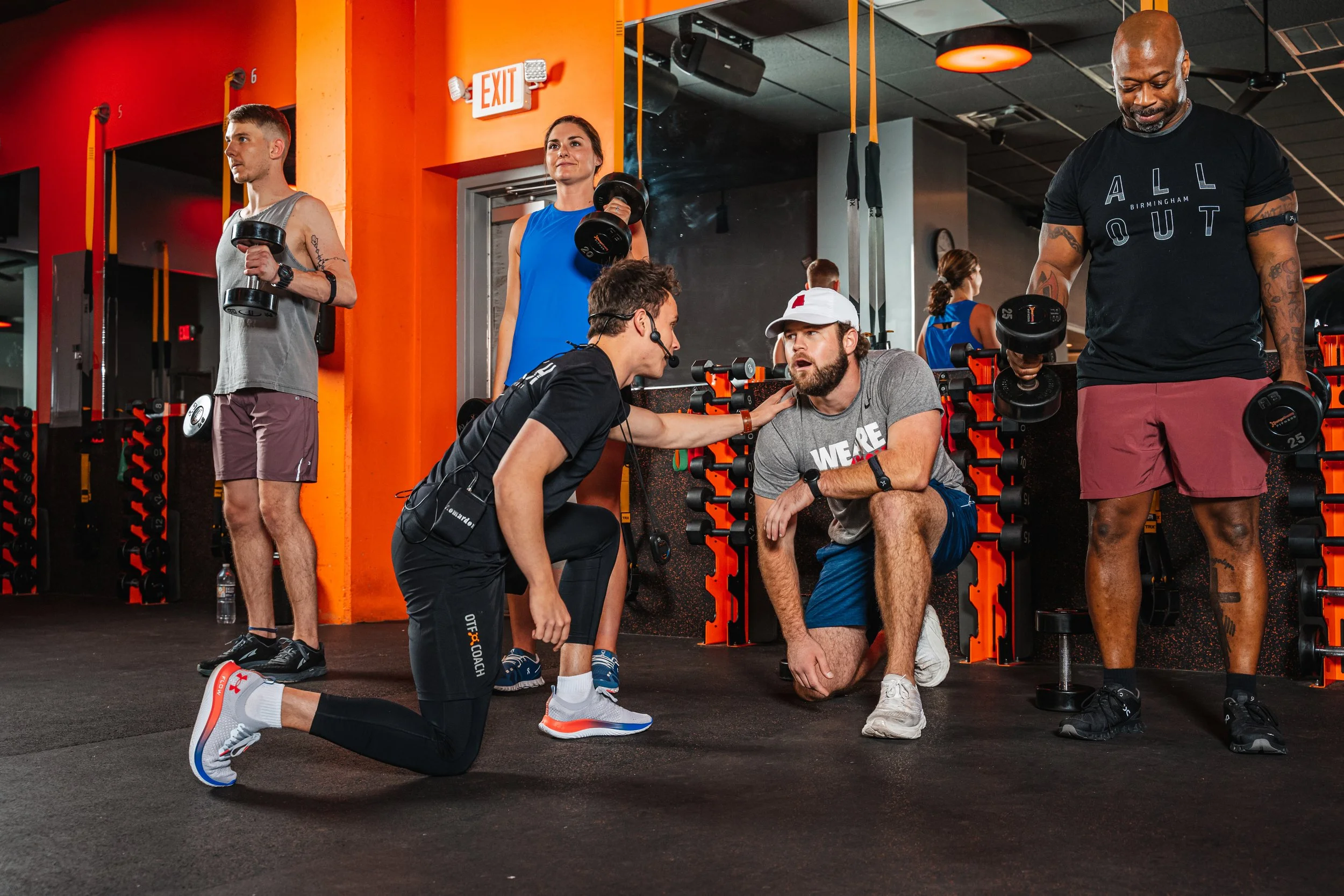 A personal trainer assists a man with a weightlifting exercise in a gym, while another person lifts dumbbells and a woman works out in the background.