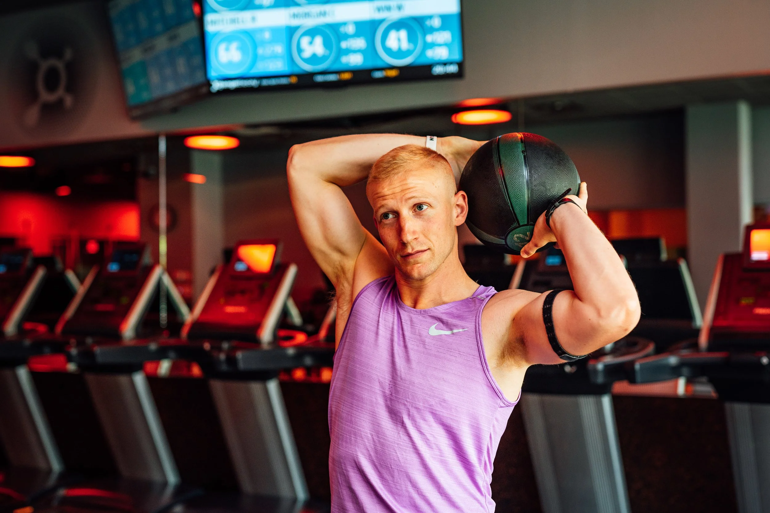 A man with a blond haircut wearing a purple workout shirt is holding a black and green medicine ball behind his head in a gym with treadmills in the background.