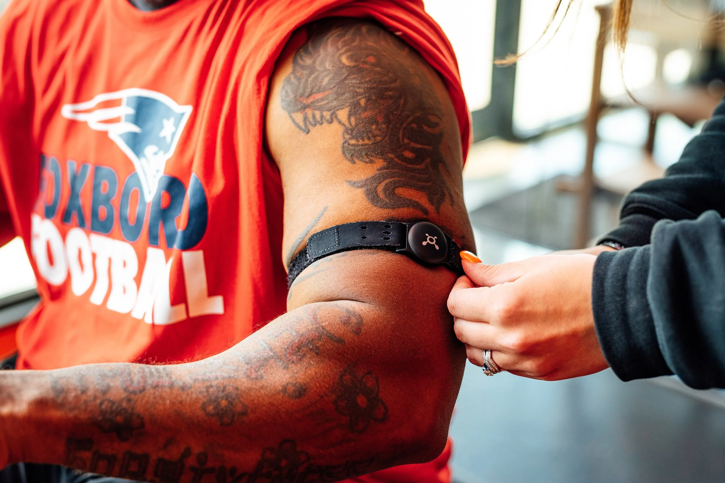 A person with a tattooed arm is assisted by a fitness coach to put on a digital bio analysis and heart beat reader band.