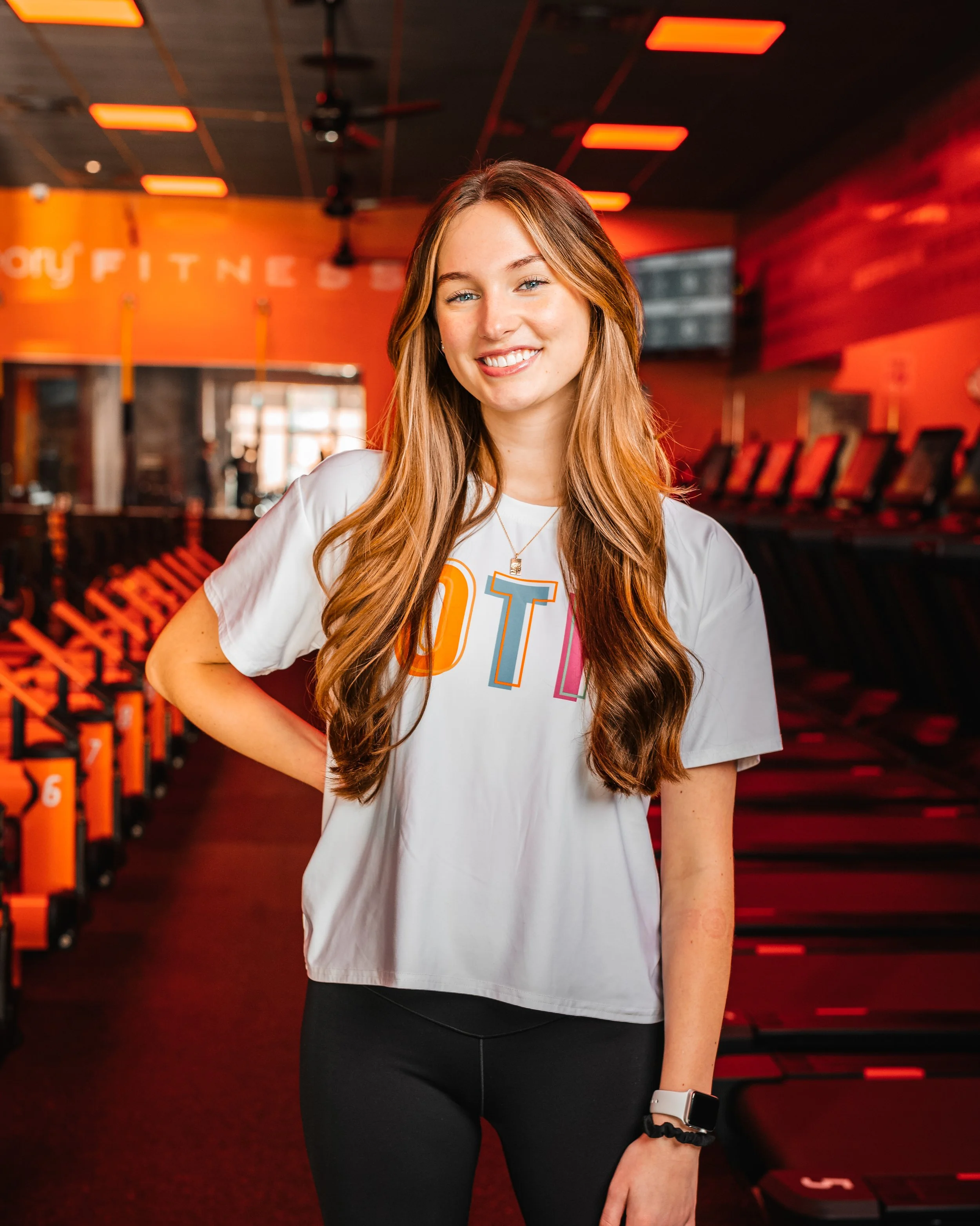A young woman with long wavy red hair, smiling, wearing a white T-shirt with "OTF" written on it, black leggings, a smartwatch, and a hair tie on her wrist, standing in a orange-lit gym with workout machines in the background.