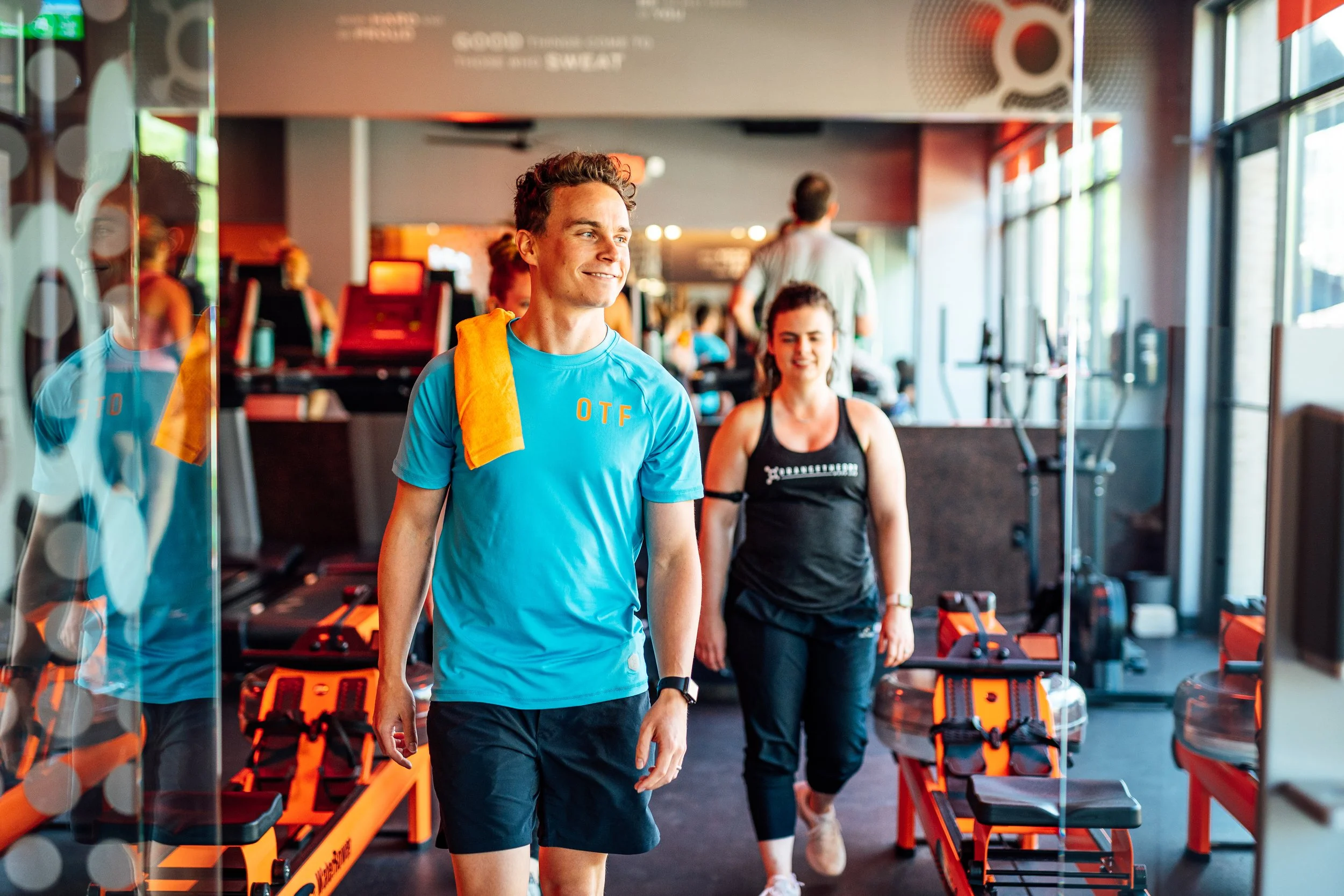 People in a gym reflecting in a glass door, two women in athletic wear, one smiling, one with a towel over her shoulder, workout equipment around.