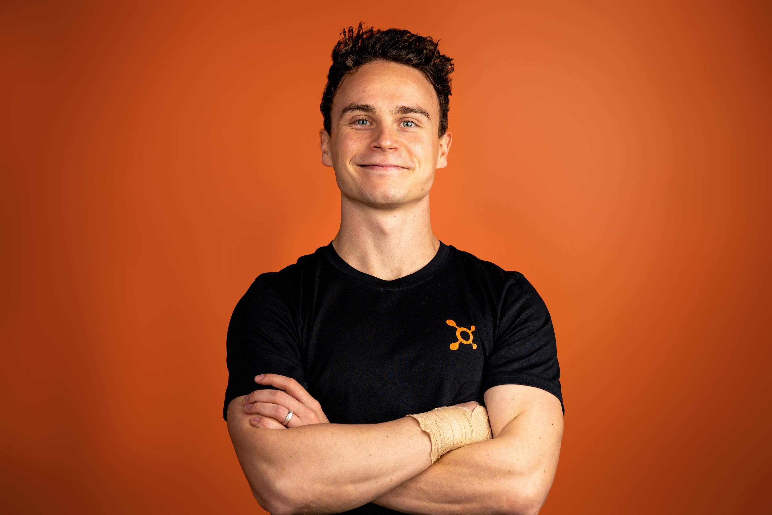 Young man smiling with arms crossed, wearing a black shirt with an Orangetheory Fitness splat logo, standing against an orange background.