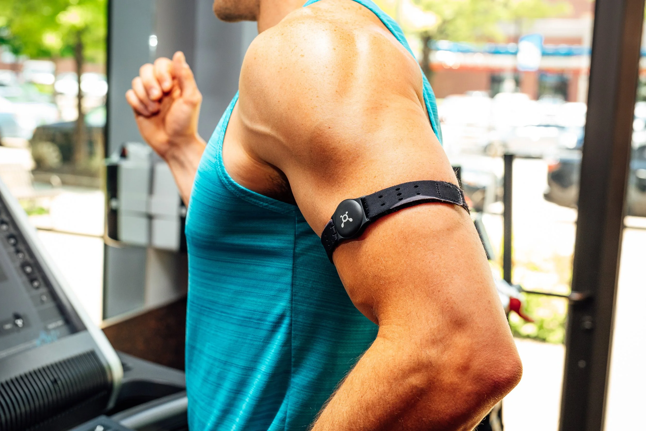 A person in a blue tank top exercising indoors on a treadmill, with a black fitness tracker on their upper arm. Large windows with a view of a street with trees and parked cars in the background.