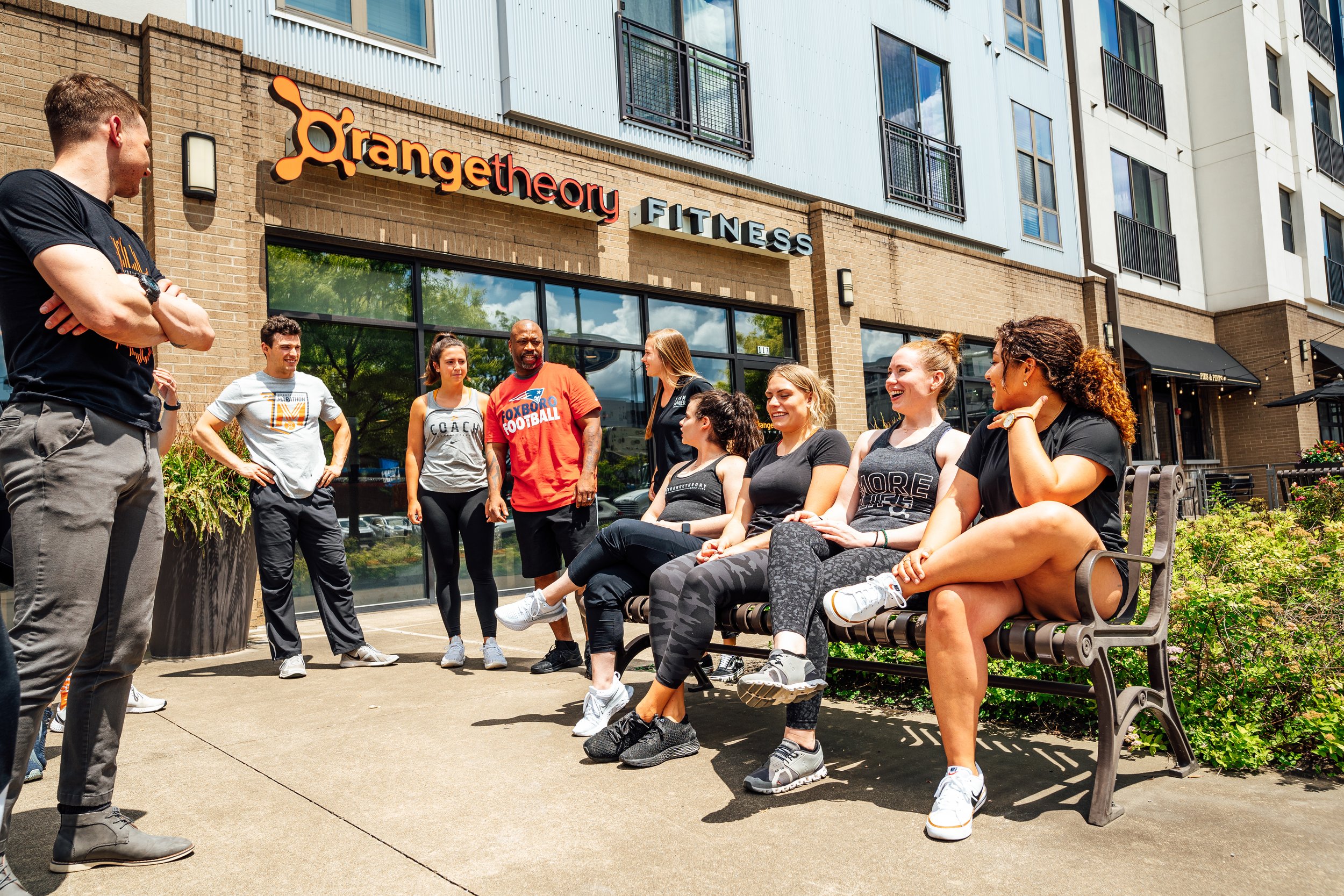 Group of people sitting and standing outside a fitness center named Orangetheory Fitness, engaging in conversation, with a man in a black t-shirt speaking to a diverse group.