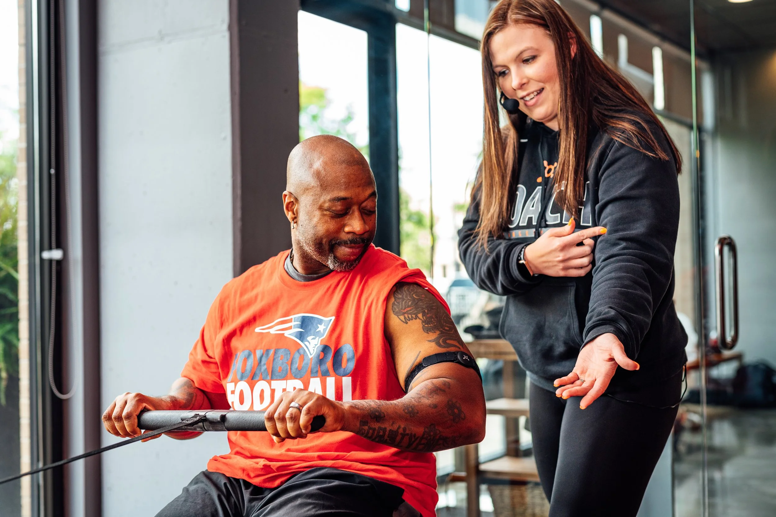 A man with a shaved head and tattoos on his arm is receiving coaching from a woman with long reddish hair and wearing a black hoodie. They are in a brightly lit indoor gym with large windows. He is seated on a rowing machine.