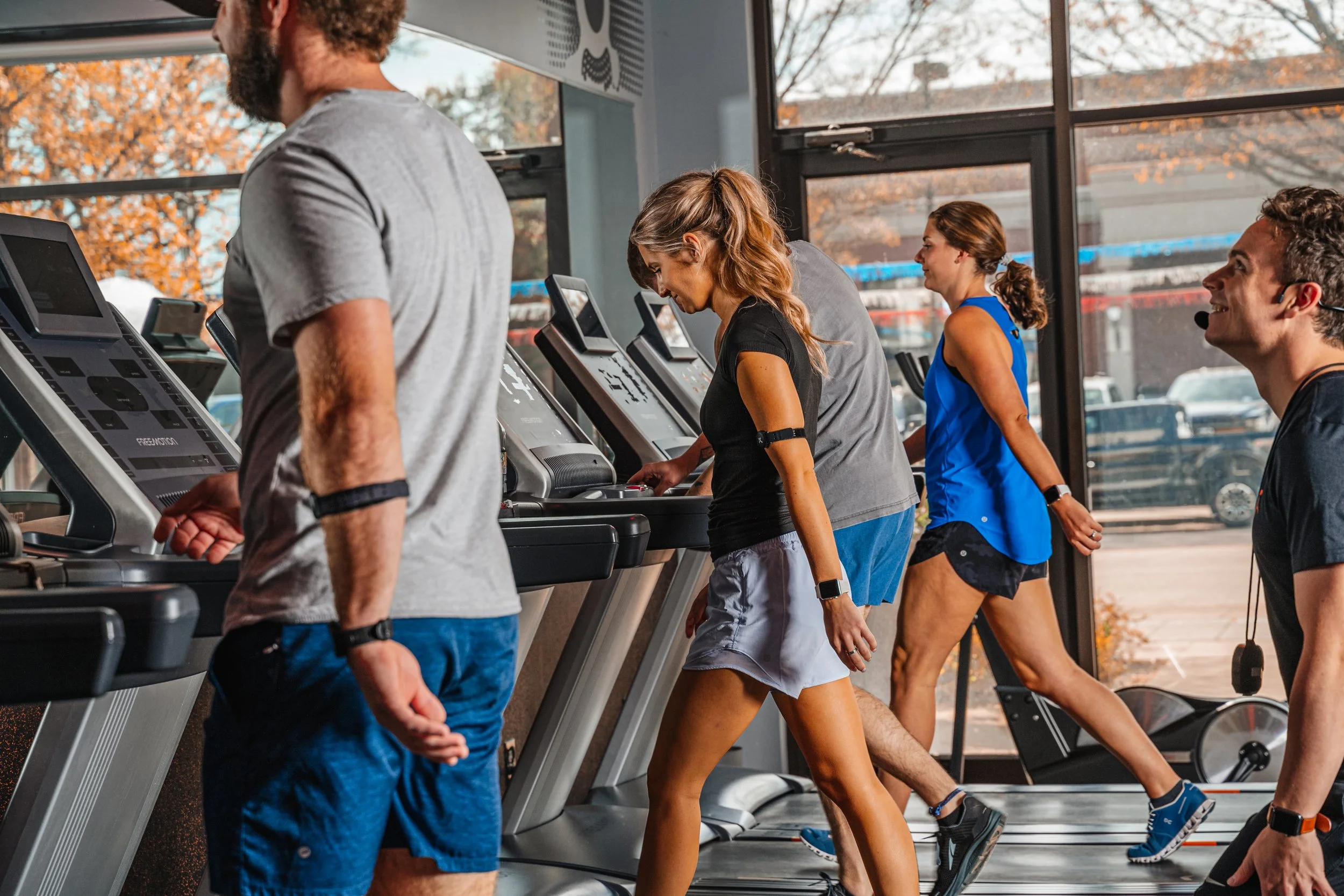People running on treadmills inside a gym, with large windows showing autumn trees outside.
