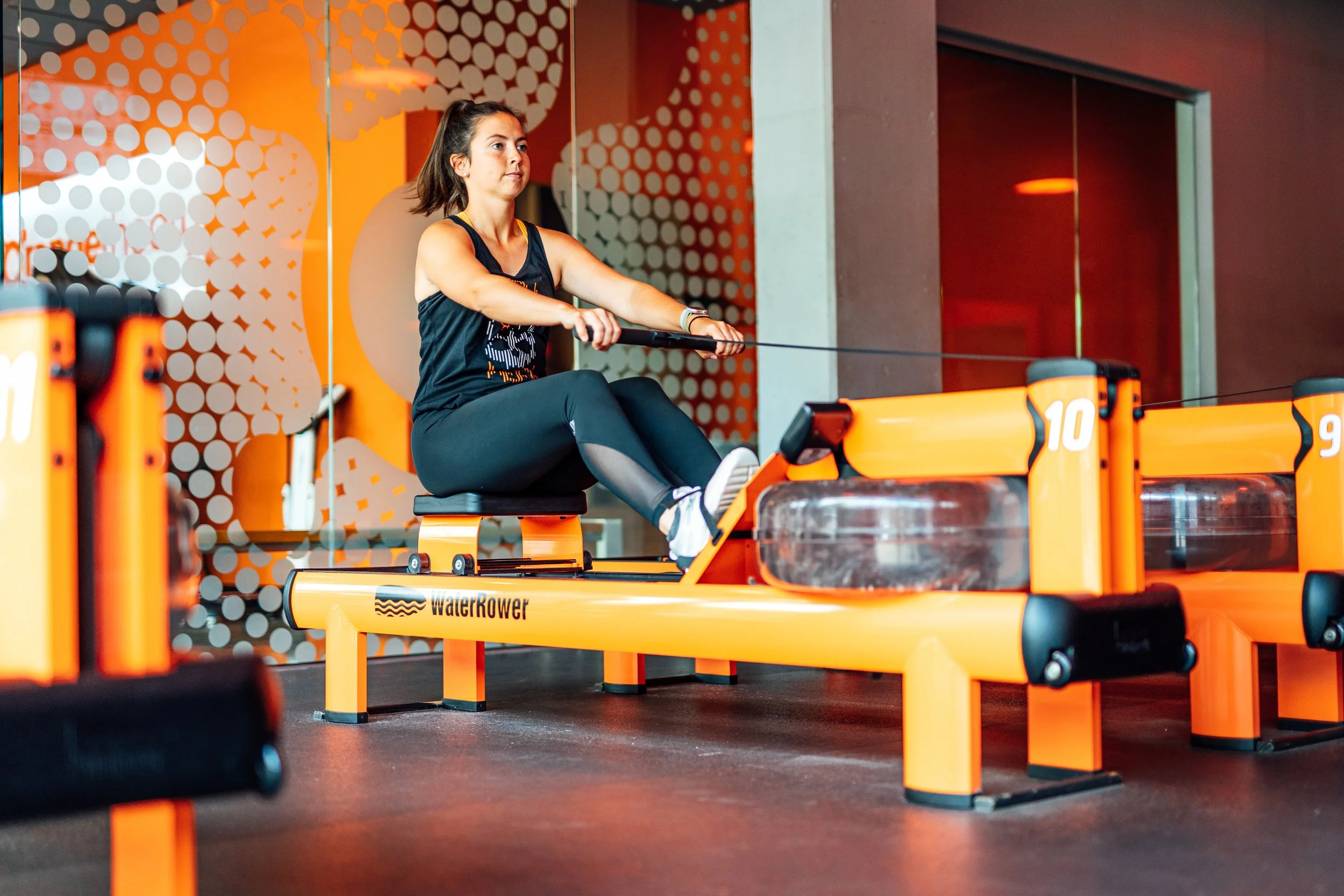 A woman performing a seated rowing exercise on an orange WaterRower indoors, with a modern orange and white dotted wall in the background.