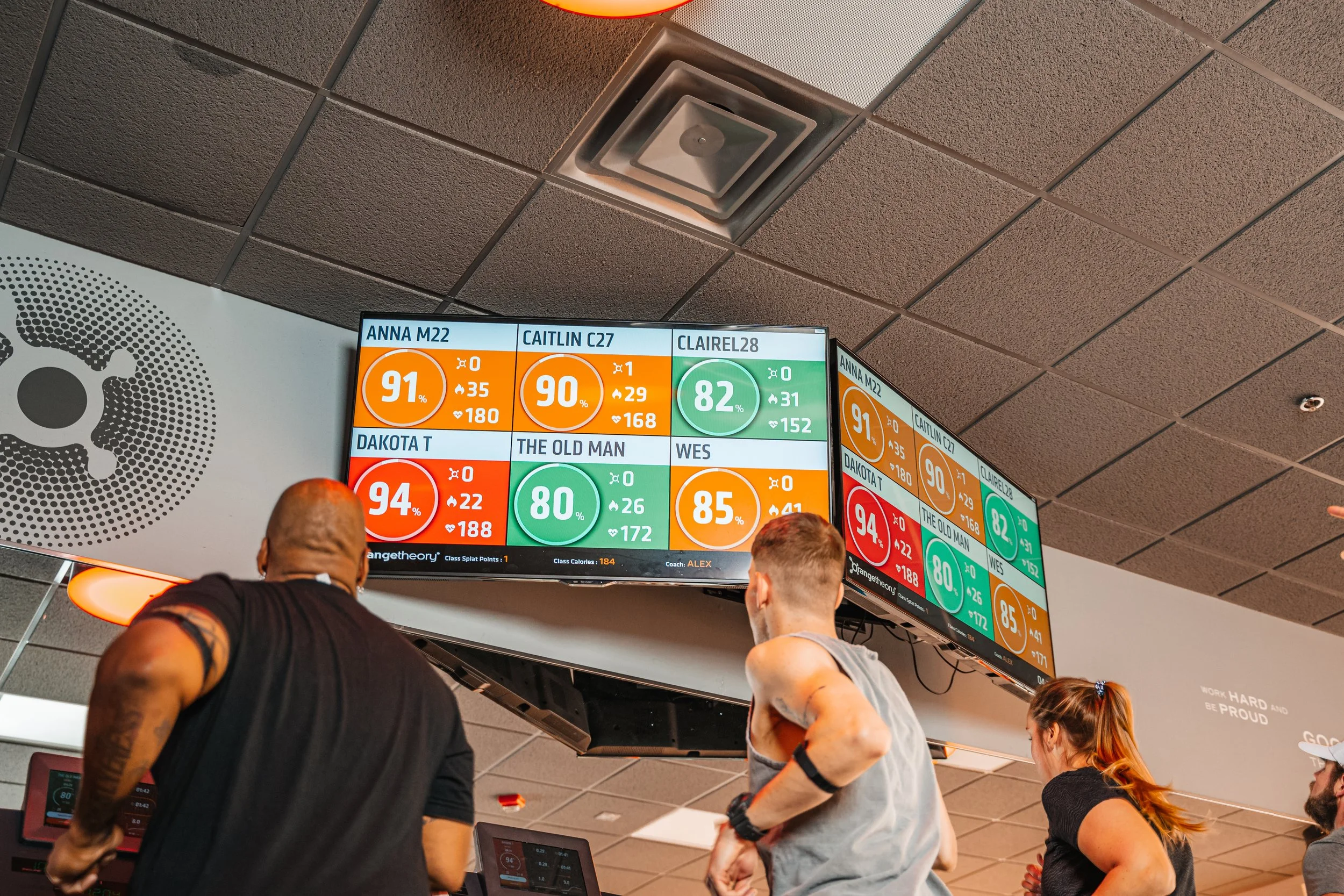 People working out on treadmills in a gym, with a digital leaderboard displaying names and scores overhead.