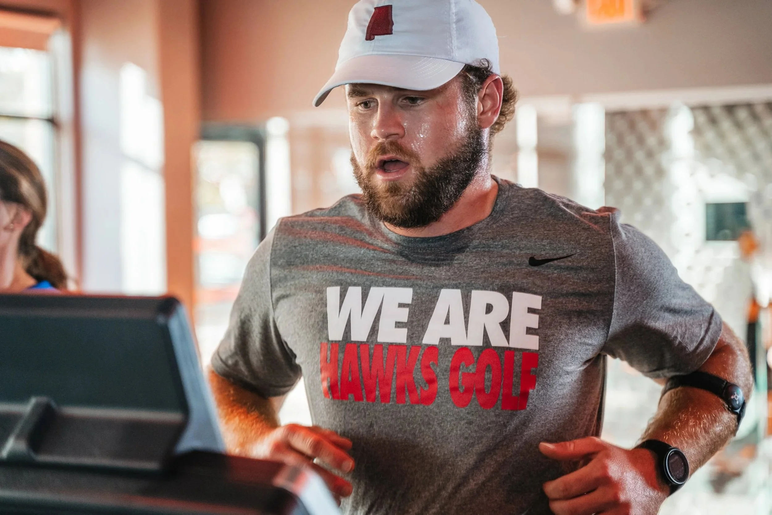 A man wearing a white cap and a gray t-shirt with red and white text, standing indoors in a gym or fitness center, looking at a monitor or screen, with a woman and other people in the background.