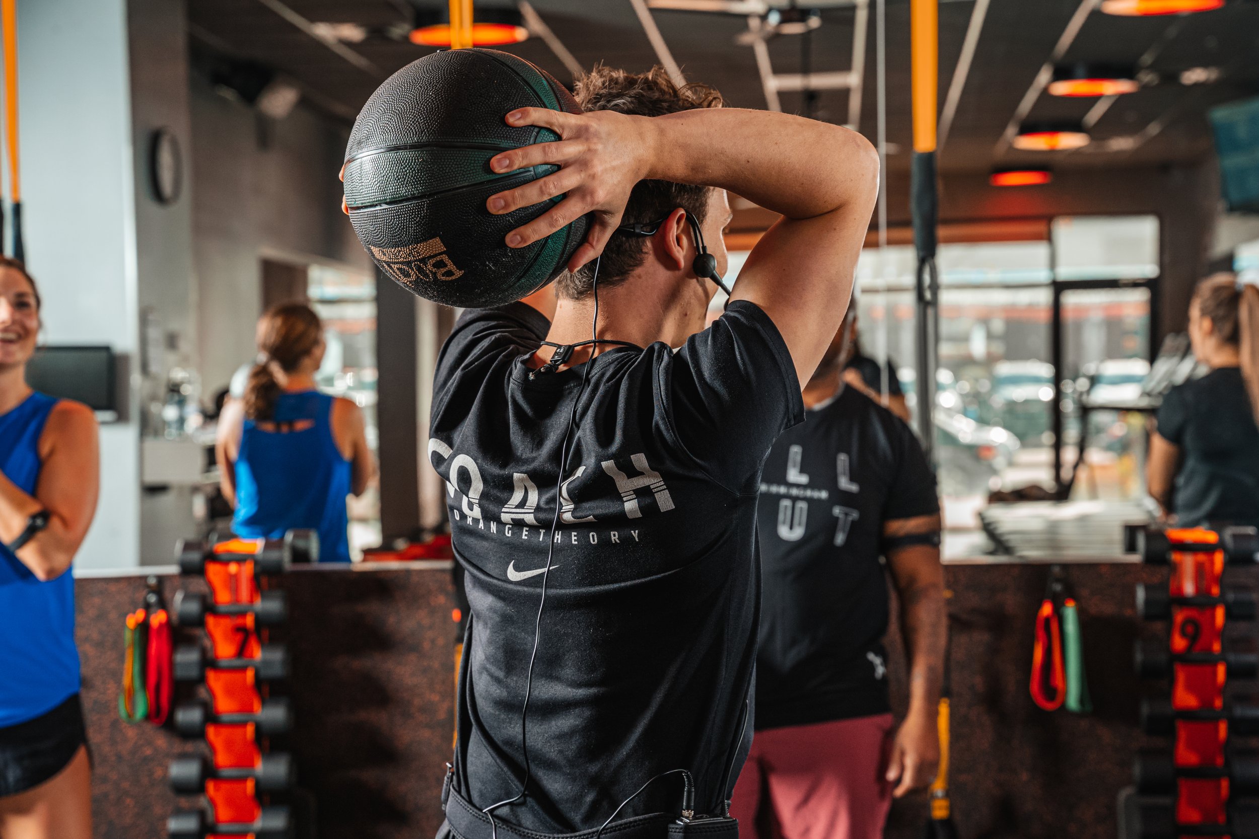 A man in a gym holding a medicine ball behind his head during a workout with other people around.