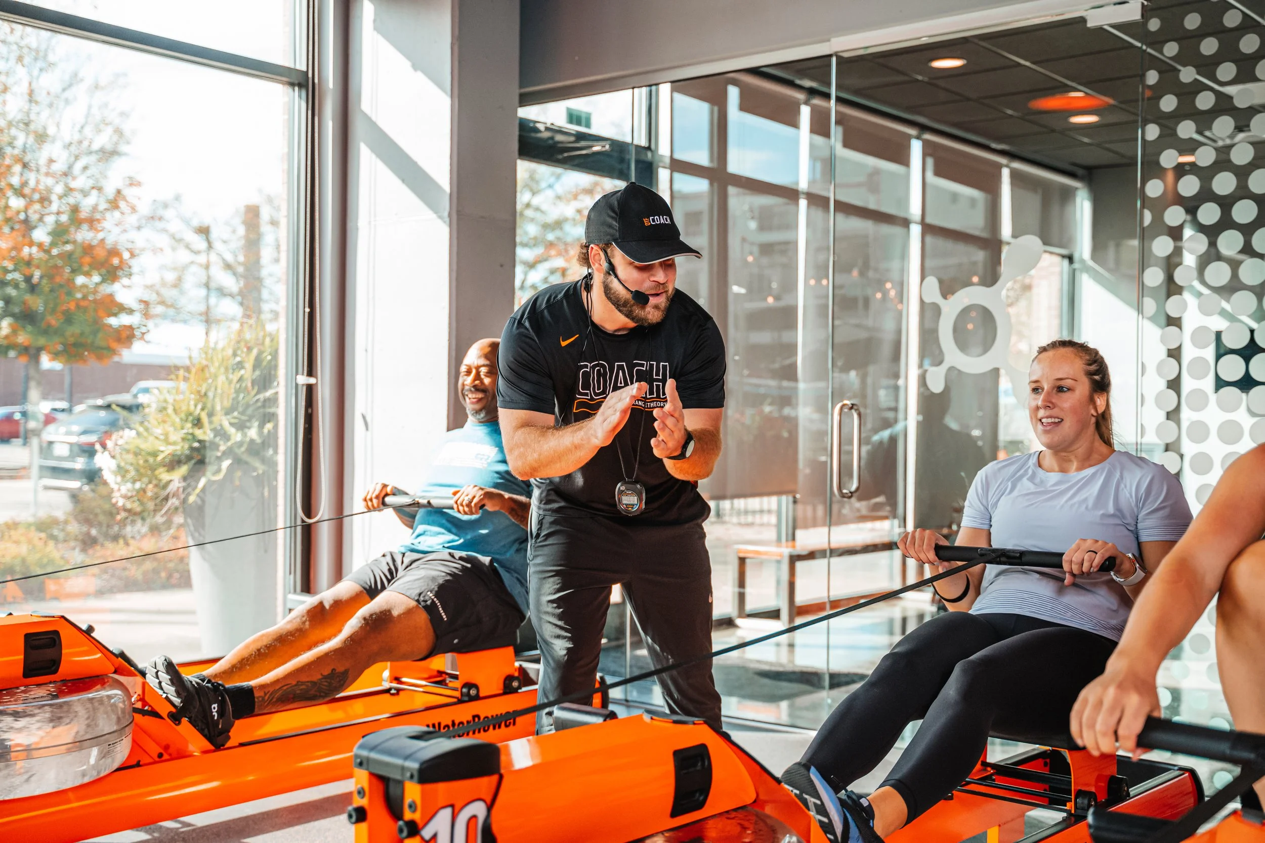 Indoor rowing workout session with a coach instructing two women on a boat ergometer machine while a man sits in the background, inside a modern gym with large windows and glass walls.