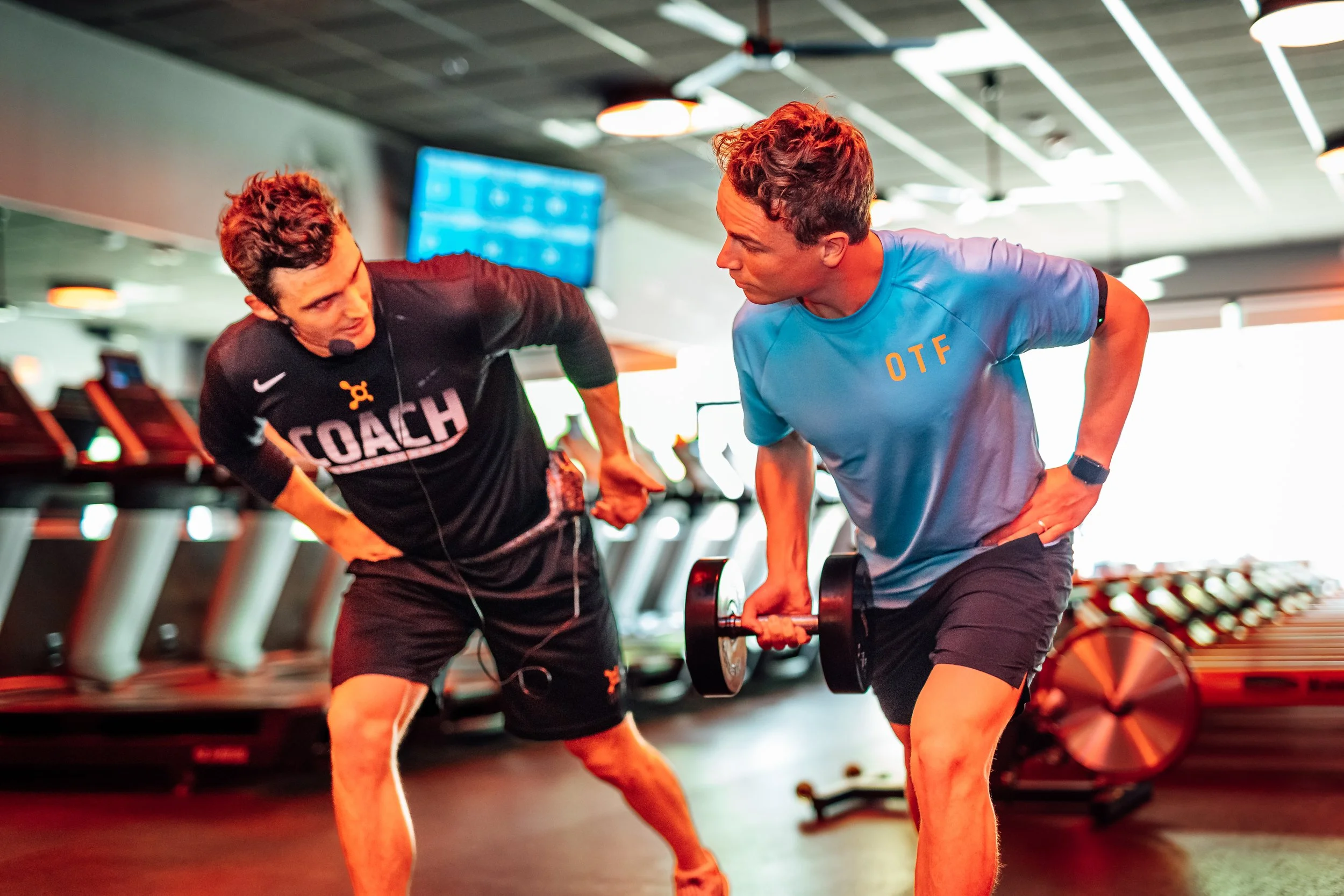 Two men involved in a workout session at a gym, with one man coaching and the other lifting a dumbbell. The gym has cardio machines in the background.