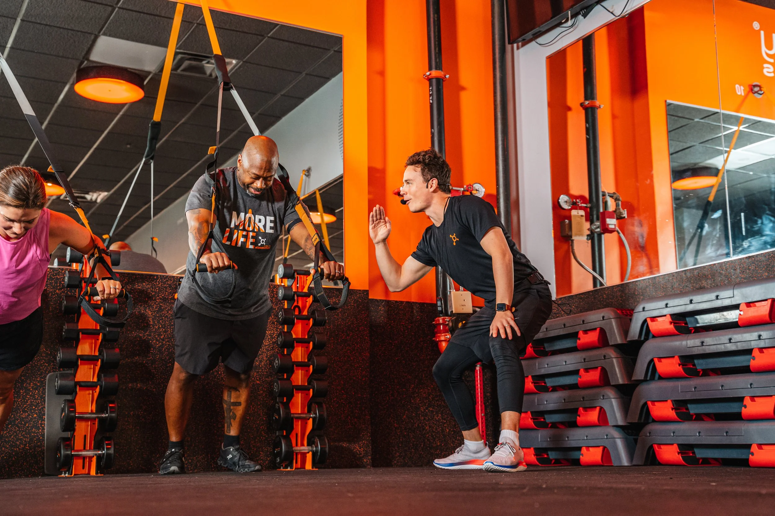 A personal trainer helping a man with suspension training straps at a gym, with a worker in black giving a high five.