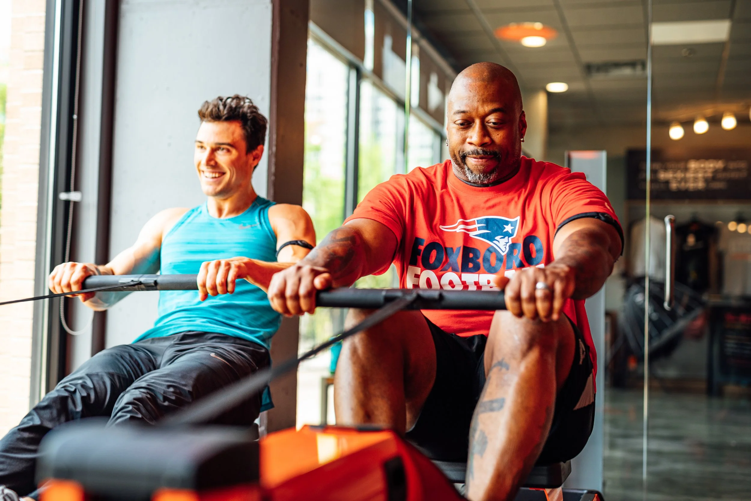 Two men are exercising on rowing machines inside a gym, smiling and enjoying their workout. The man in the foreground wears a red t-shirt with a sports team logo, and the man in the background wears a blue tank top.