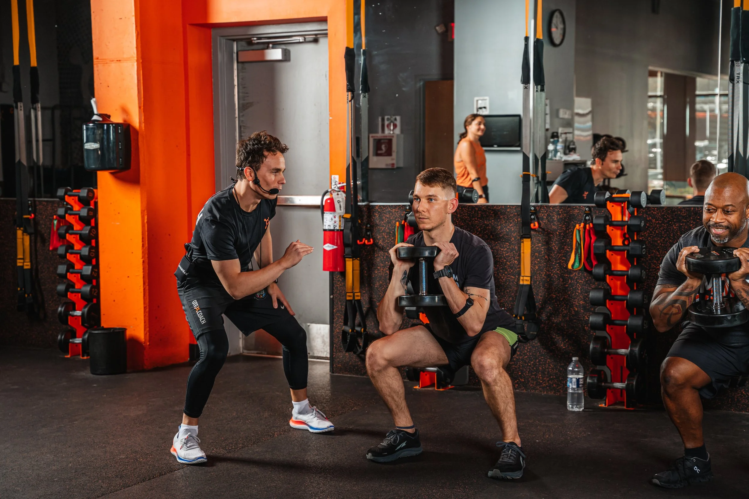 A fitness trainer instructs a man in a gym with workout equipment and other people exercising in the background.