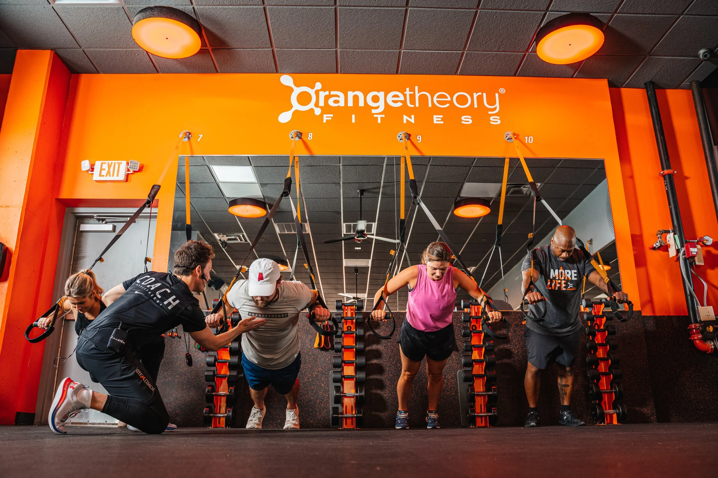 People participating in a workout class at Orangetheory Fitness, using suspension training equipment with orange and black accents.