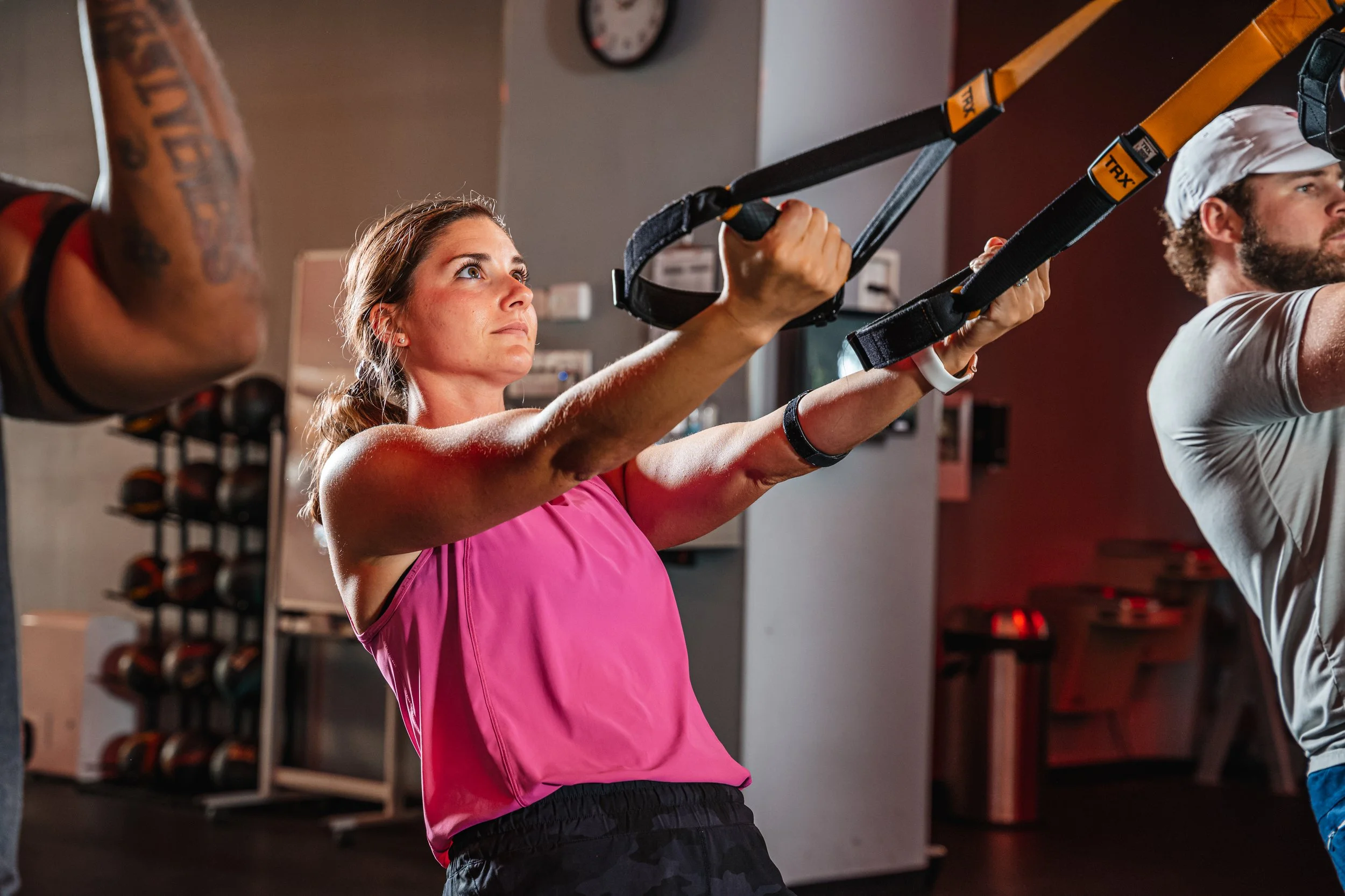 A woman in a pink tank top and black shorts is using TRX suspension trainers for workout in a gym. She is focused and pulling on the straps. A man in a white cap and shirt is exercising beside her. Gym equipment, including weight racks, is visible in the background.