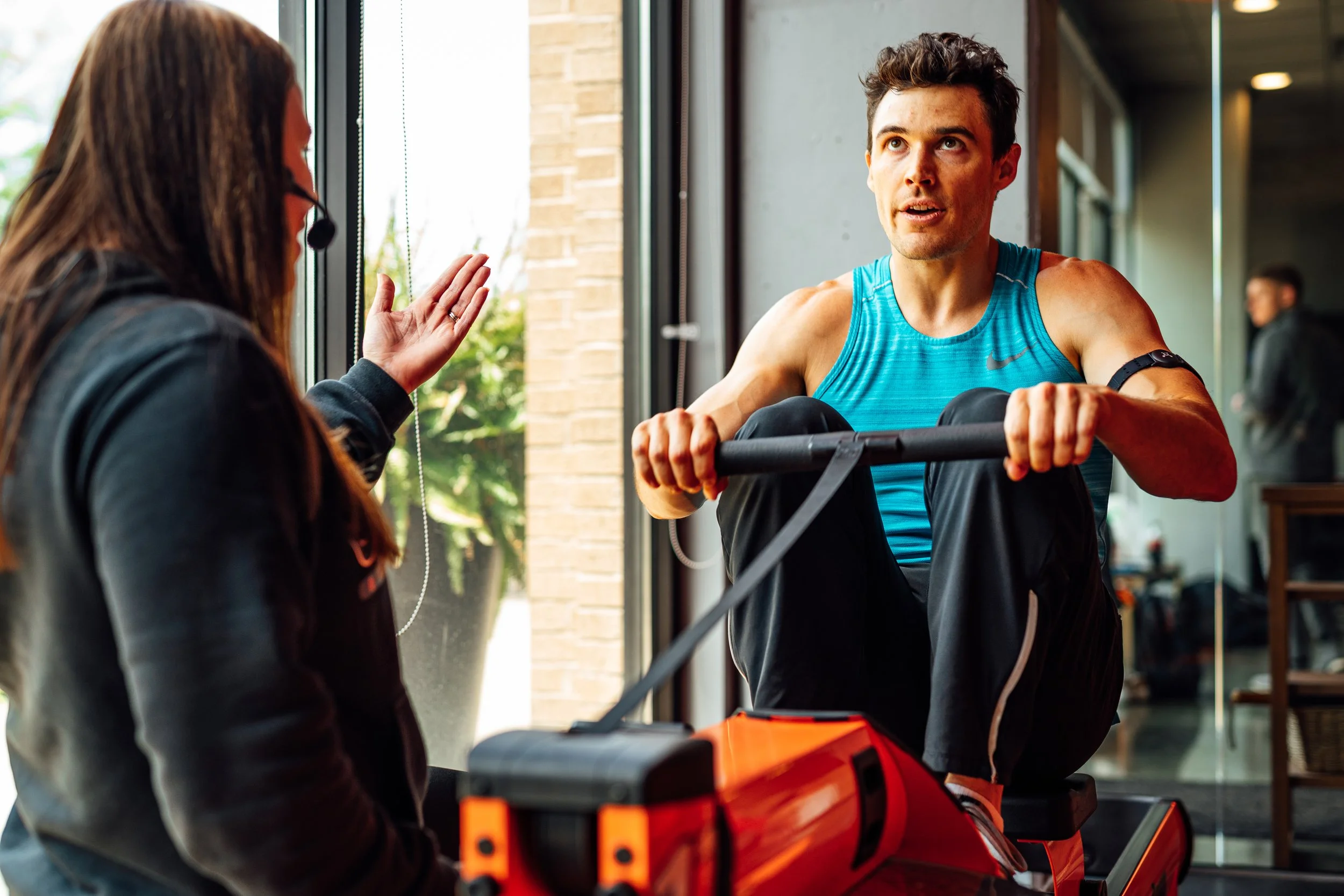 A man in a blue athletic shirt sitting on a wooden bench and talking to a woman with a headset near a large window in a gym or fitness center.