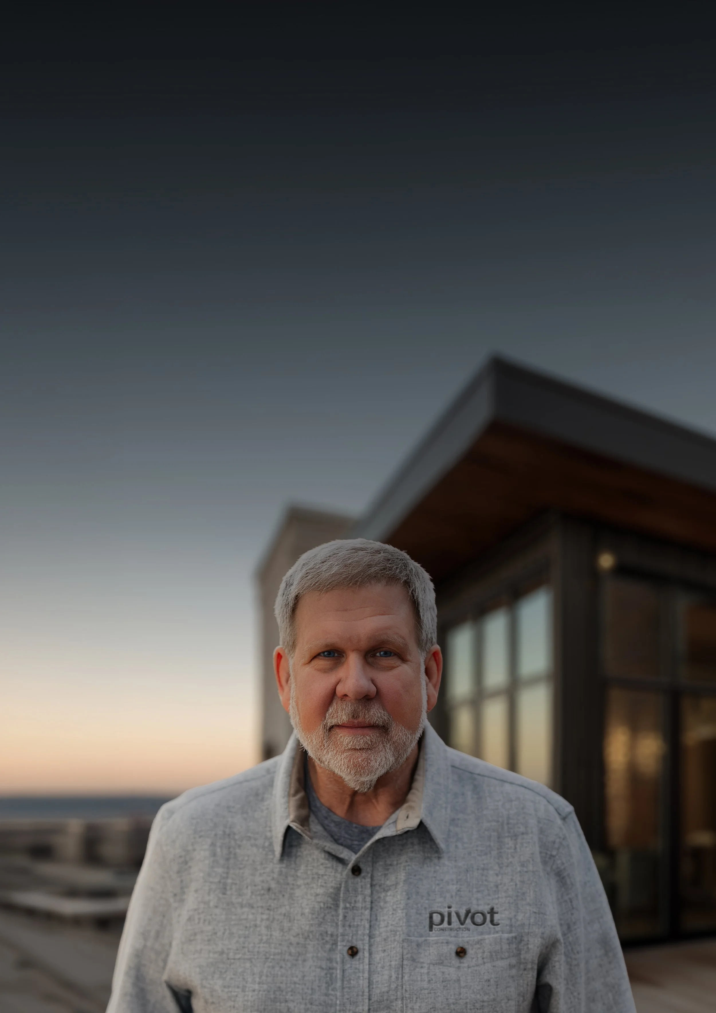 A portrait Mark Holliday, founder of Pivot Construction, wearing a gray collared shirt. He is standing outdoors during sunset, with a modern building with large windows in the background.