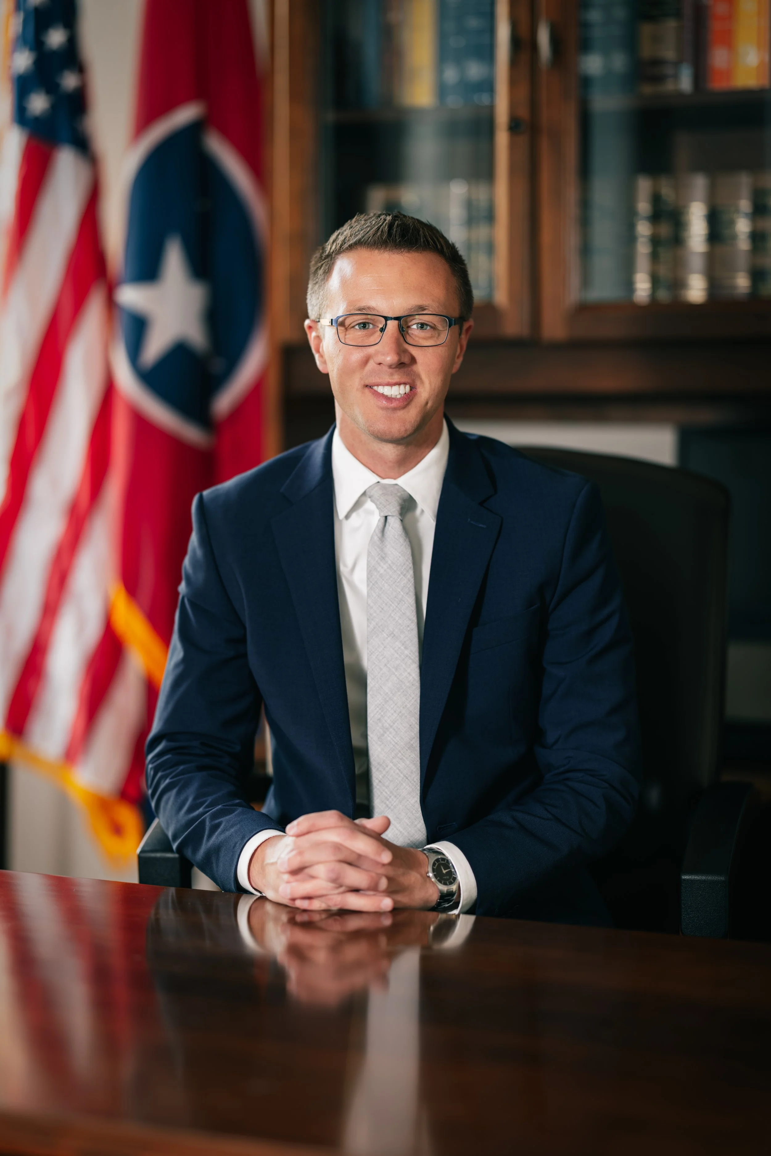 A man in a navy blue suit, white shirt, and silver tie sitting at a wooden conference table with hands clasped, smiling, in a formal office setting with an American flag and a Tennessee state flag in the background, behind glass-fronted bookshelves.