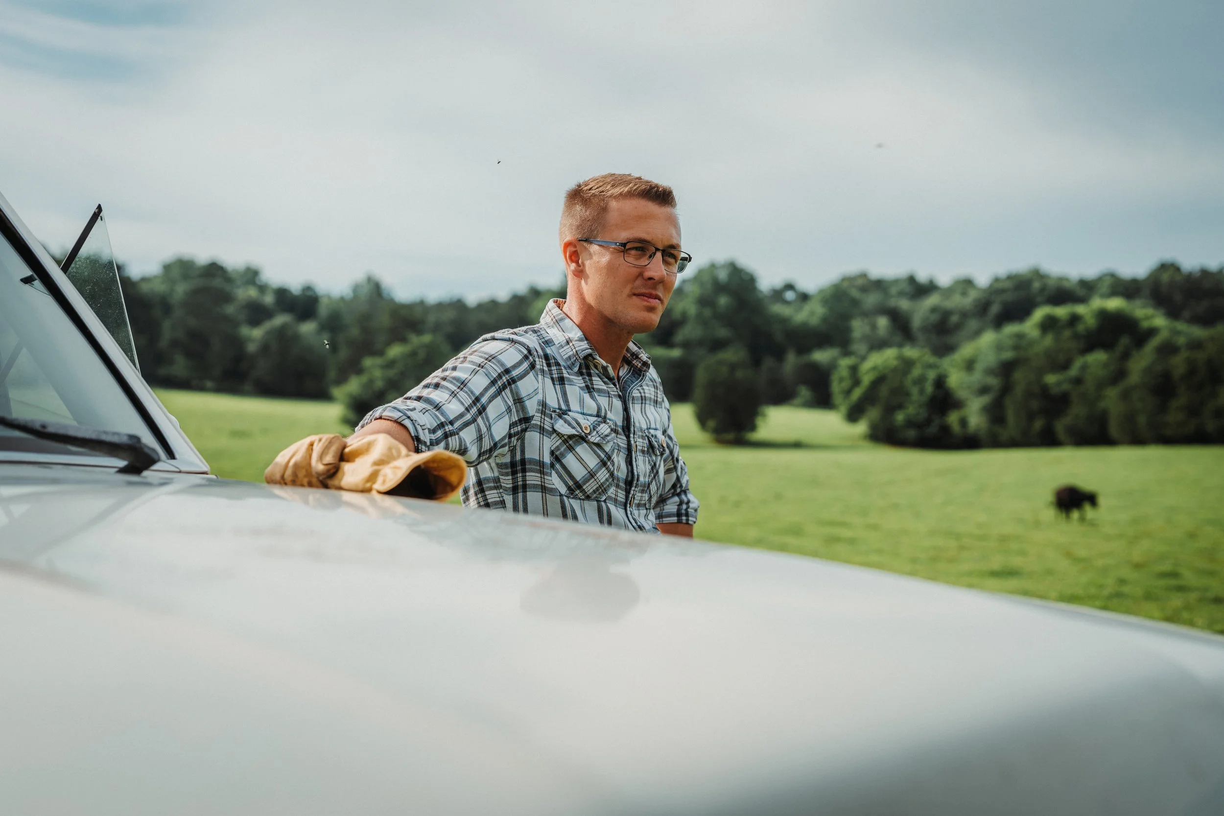 A man with glasses and a plaid shirt standing outdoors near a vehicle in a green field, with trees and cloudy sky in the background.