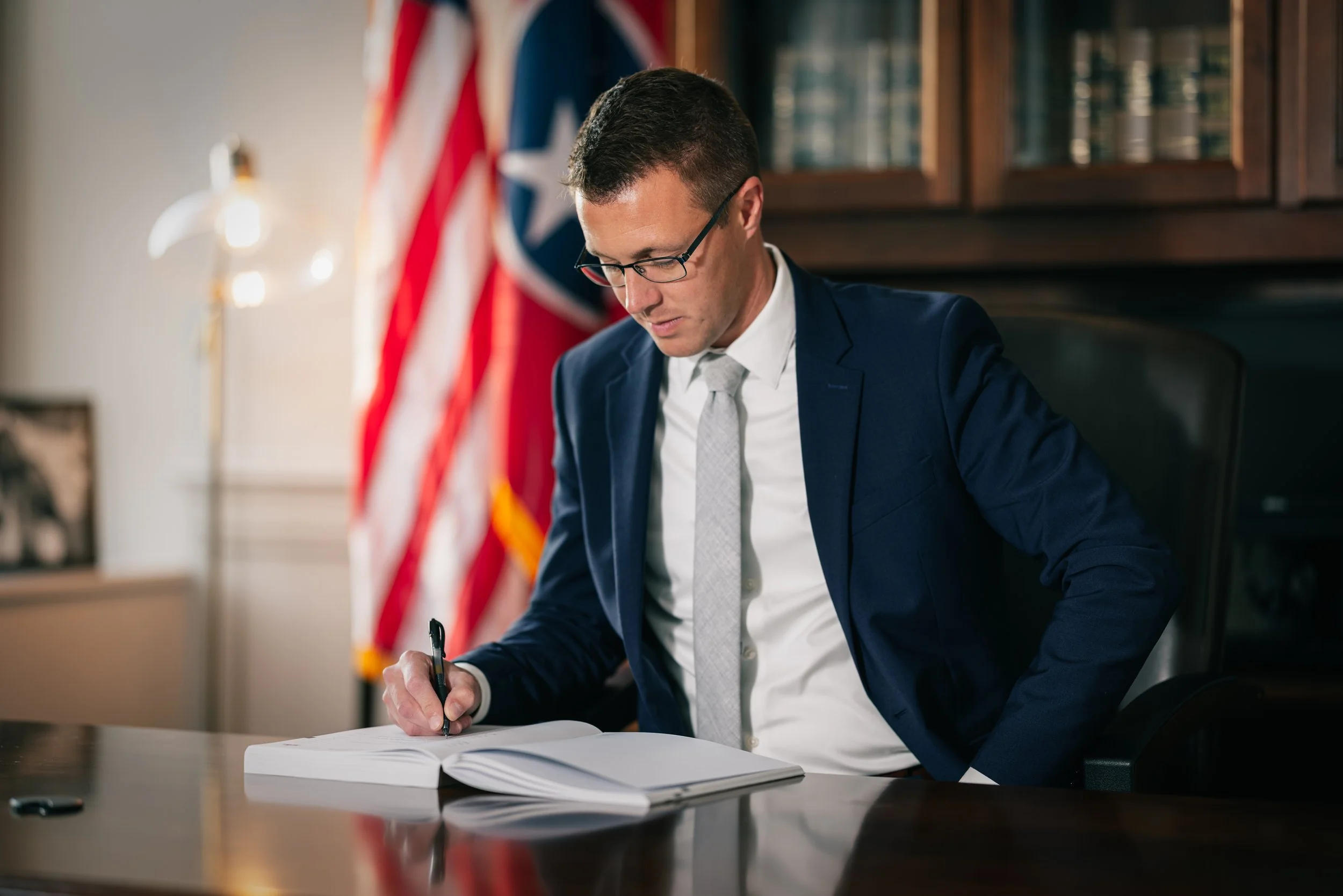 A man in a blue suit with glasses sitting at a desk, signing a document. Behind him, there are American flags and a wooden bookshelf with glass doors.