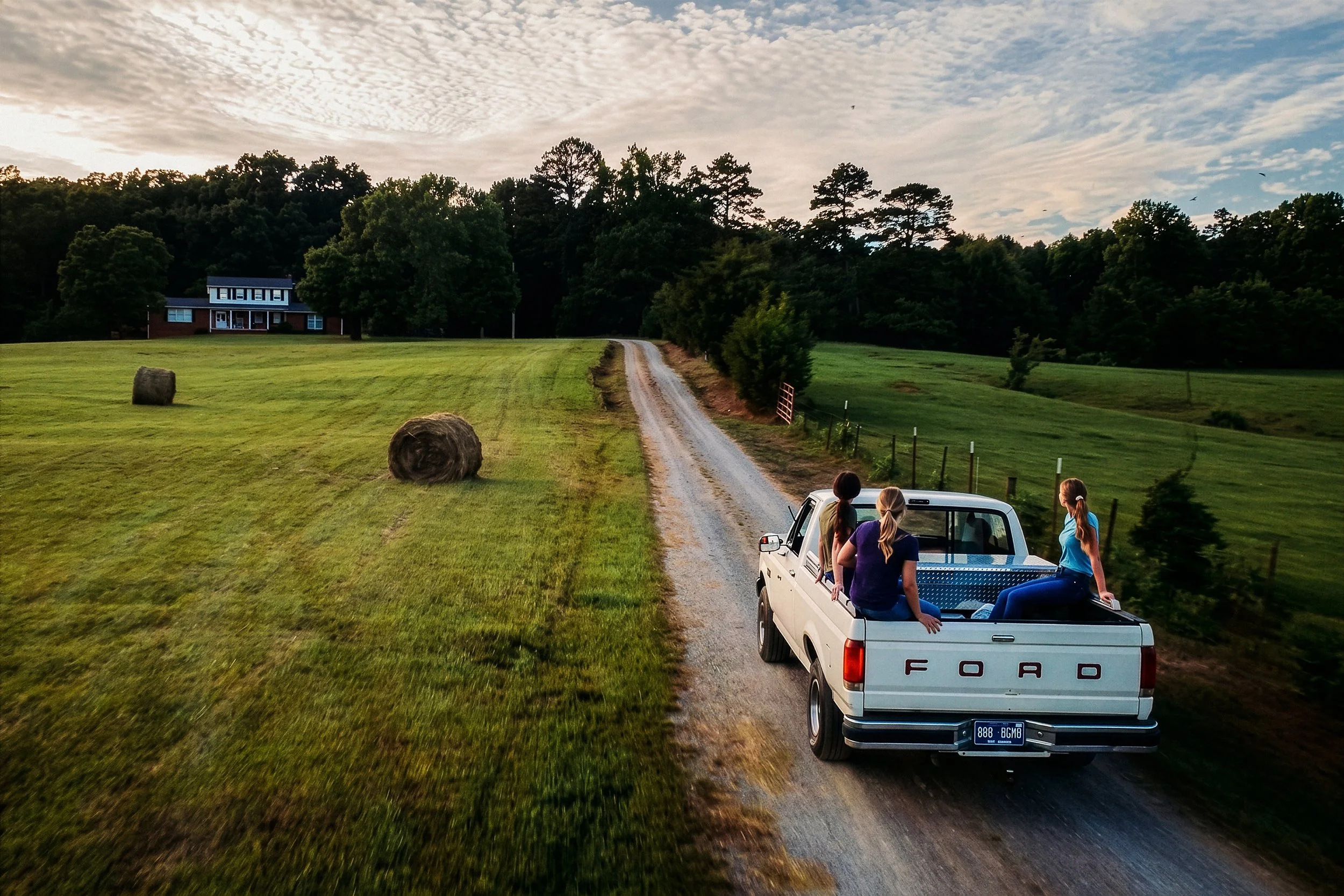 A white Ford pickup truck driving on a dirt road in a rural area with three women sitting in the truck bed, overlooking a lush green field with hay bales. There is a house in the distance, trees lining the horizon, and a partly cloudy sky overhead.