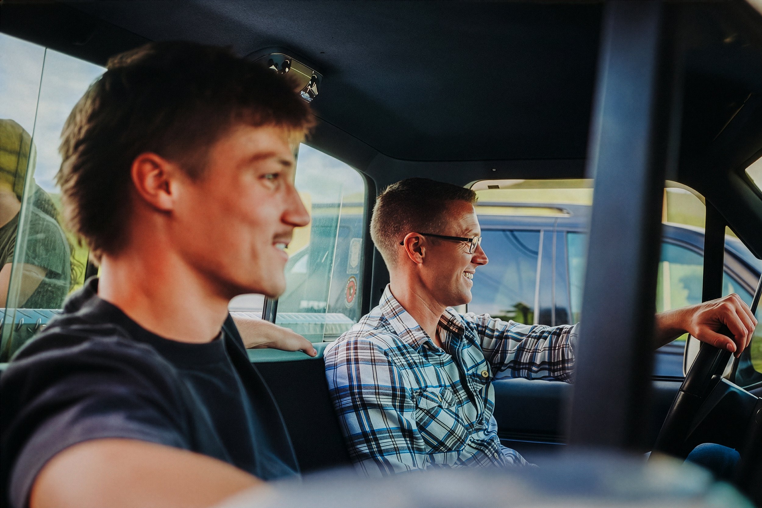 Two young men sitting in the front seats of a vehicle, smiling and enjoying the ride under a blue sky with clouds.