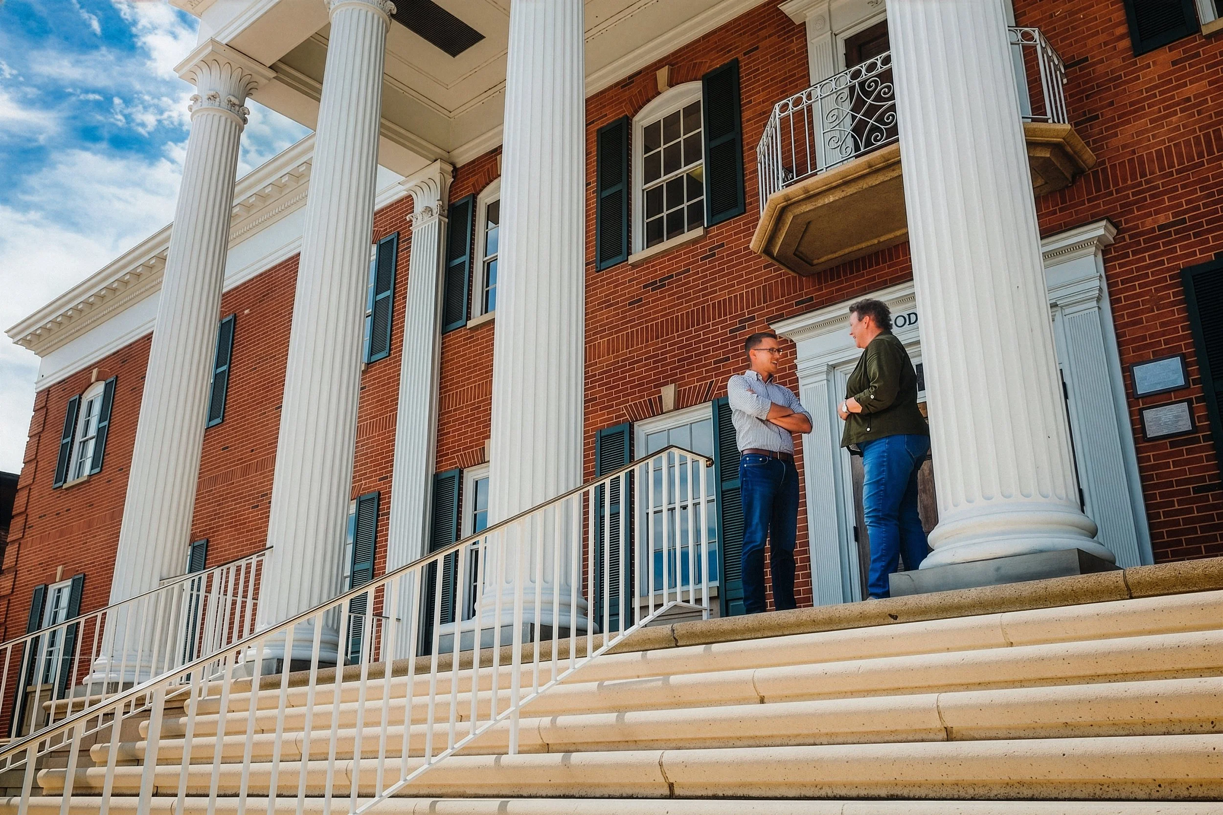 Two people stand on the steps of a large, brick mansion with white columns and black shutters, engaged in conversation under a partly cloudy sky.