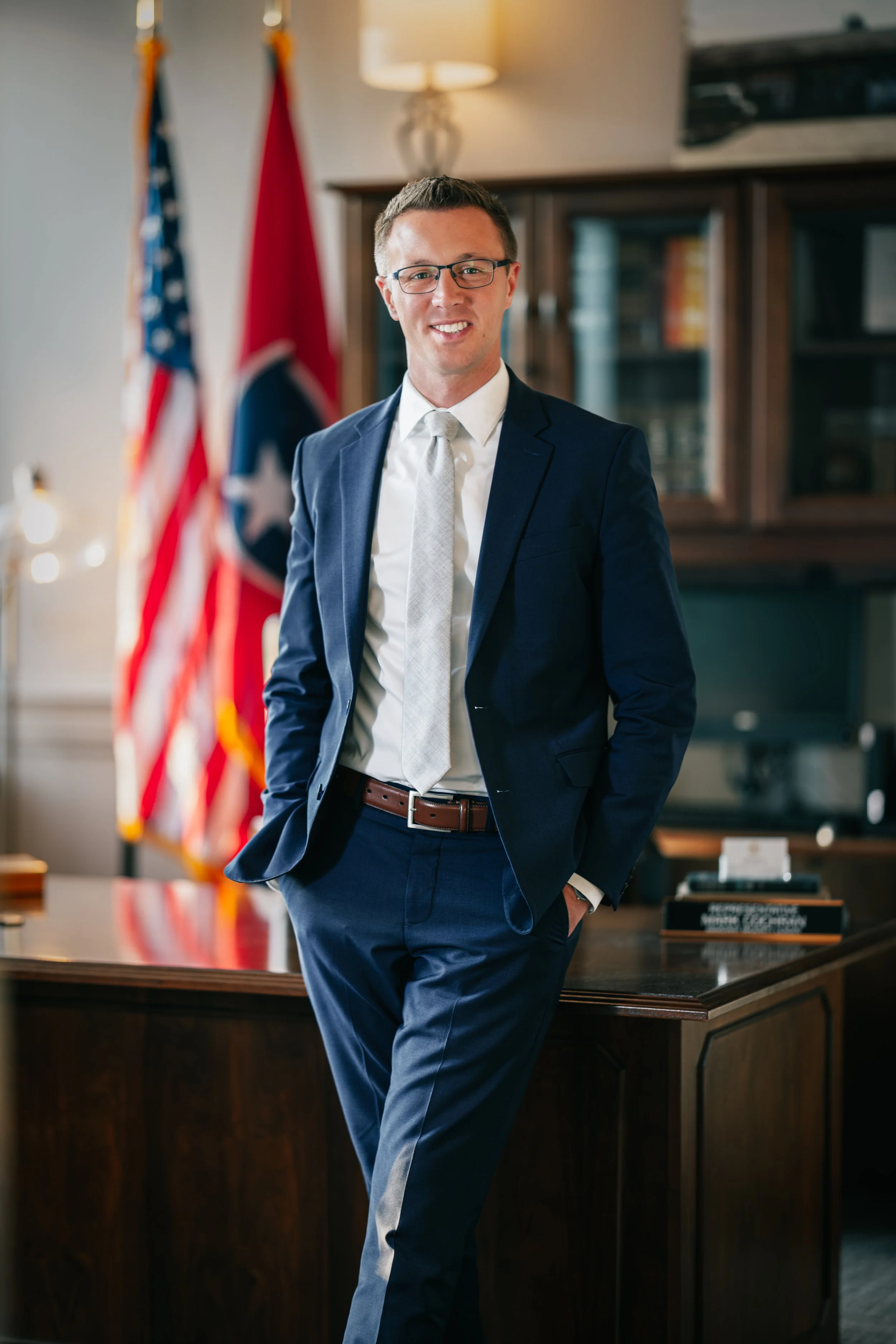 A young man in a navy suit, white shirt, and light tie standing in an office with flags in the background, smiling and leaning against a wooden desk.