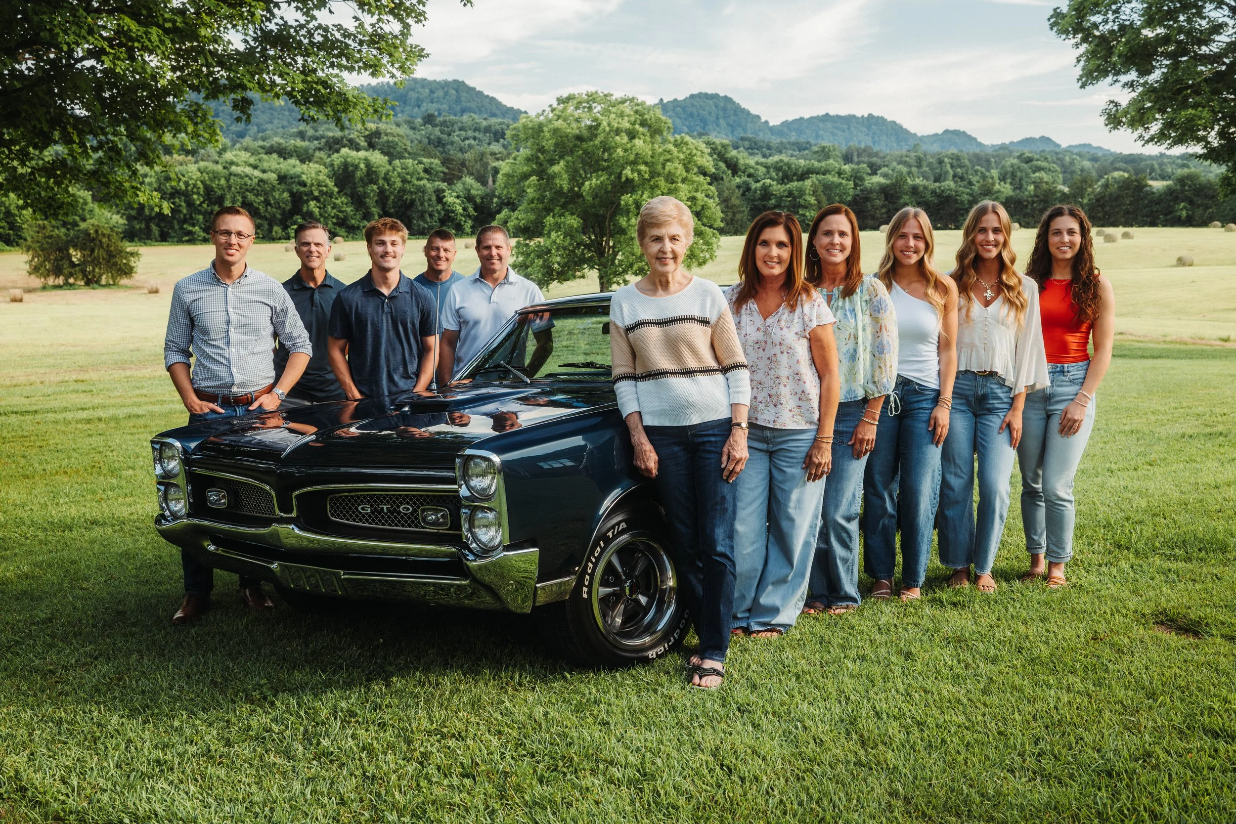 A family of eleven people standing together outdoors beside a black vintage muscle car on a grassy field with trees and mountains in the background.