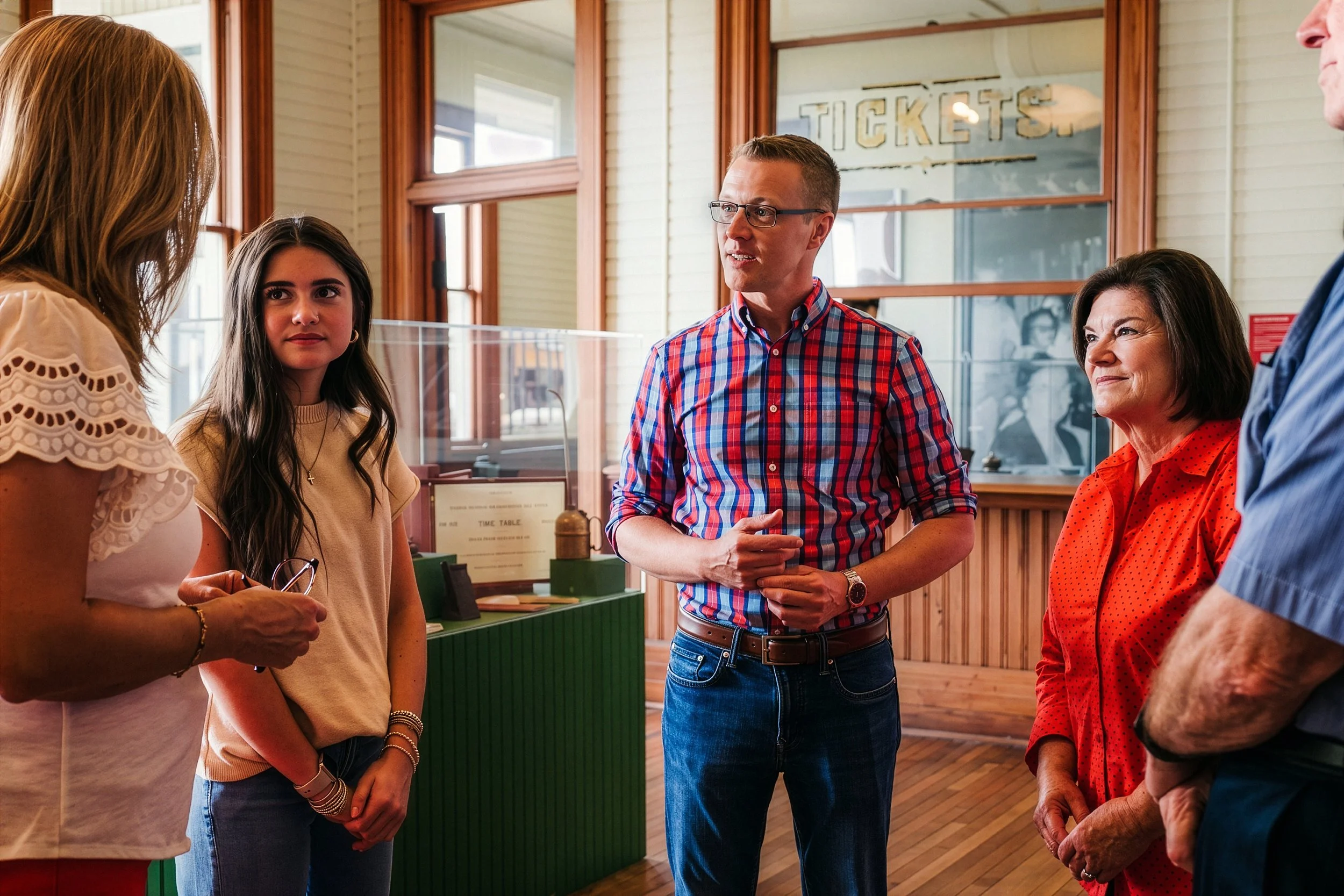Group of five people talking in a ticket booth area with wood-paneled walls. One woman in a white lace top is talking, while a young woman in beige listens attentively. A man in a red plaid shirt is speaking, and an older woman in red polka dots listens. Part of another person is visible on the right.