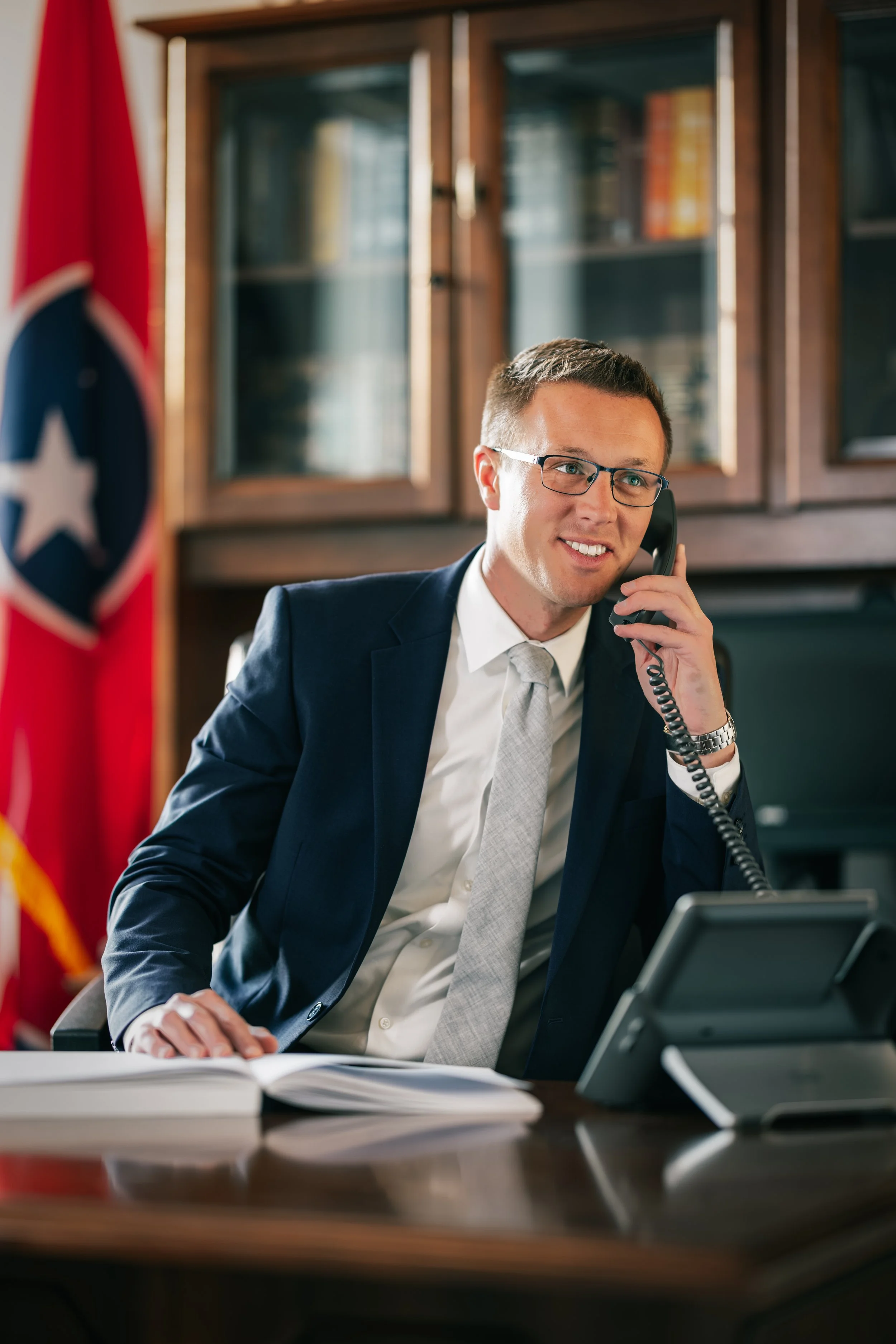 A man wearing glasses, a dark suit, and a tie talking on a landline phone at an office desk. There is an American flag and a wooden bookcase with books in the background.