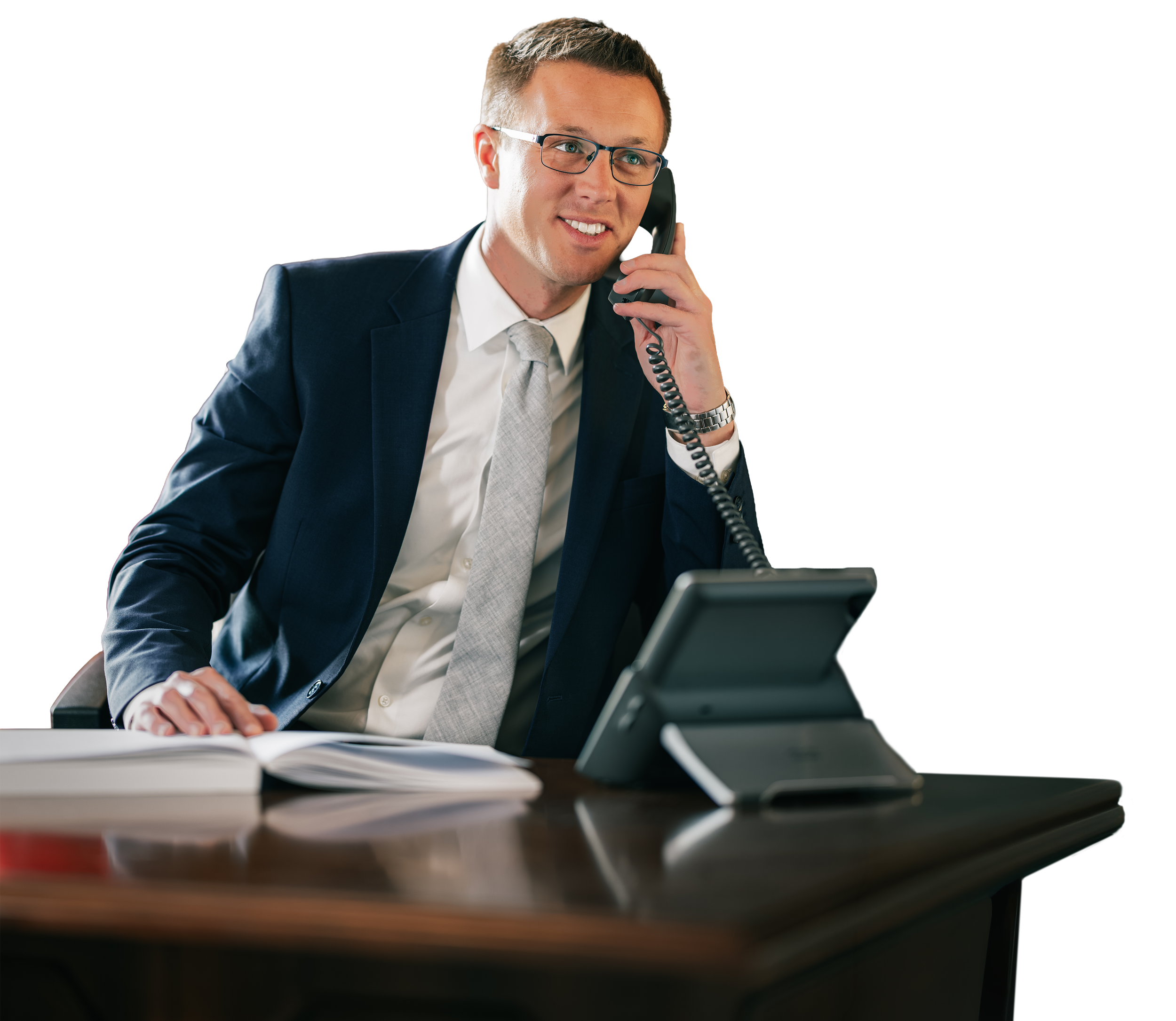 A man wearing glasses in a business suit talking on a landline phone at an office desk with open notebook and a computer monitor.