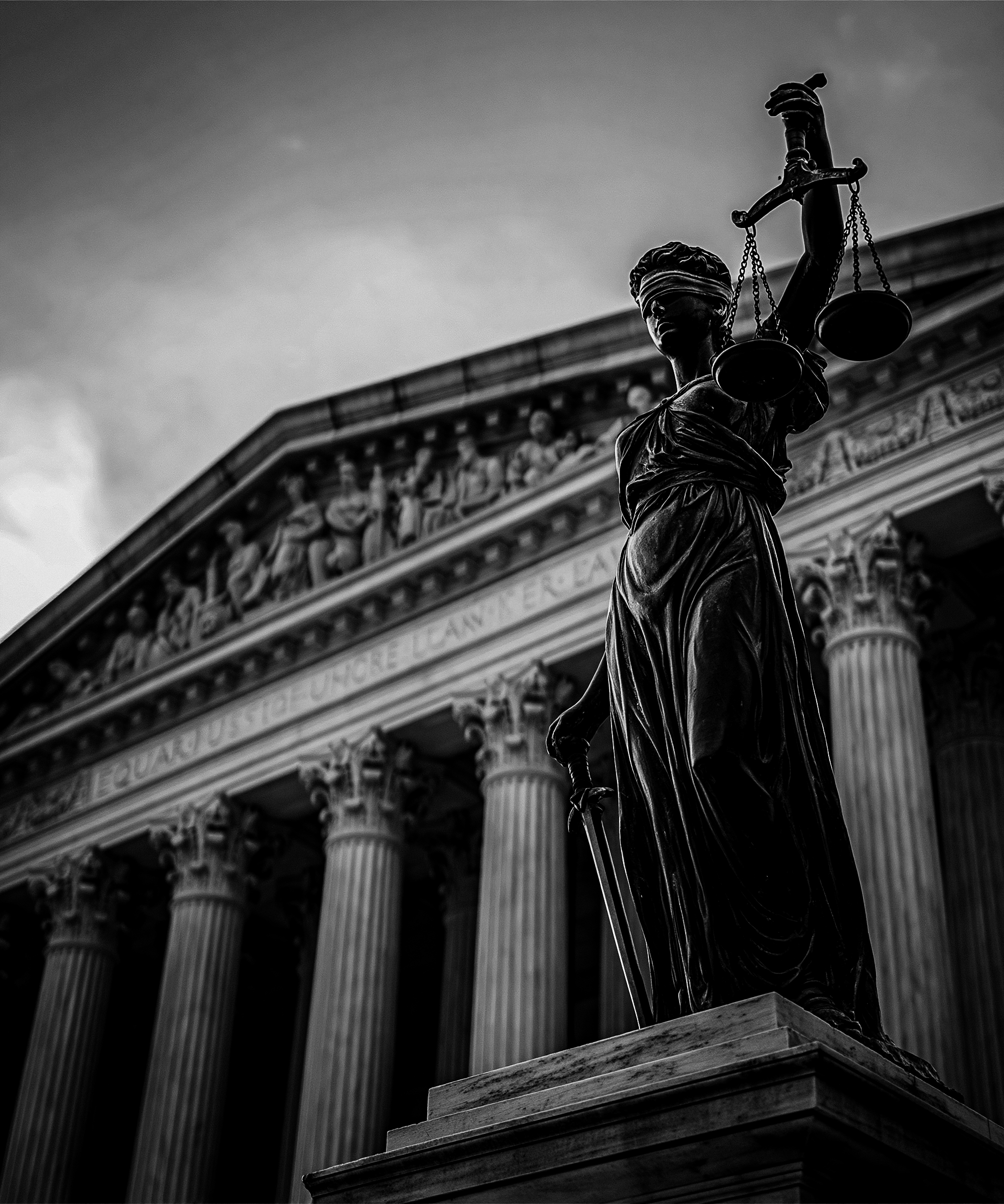Black and white photograph of the statue of Lady Justice holding scales in front of a classical building with columns and intricate carvings.