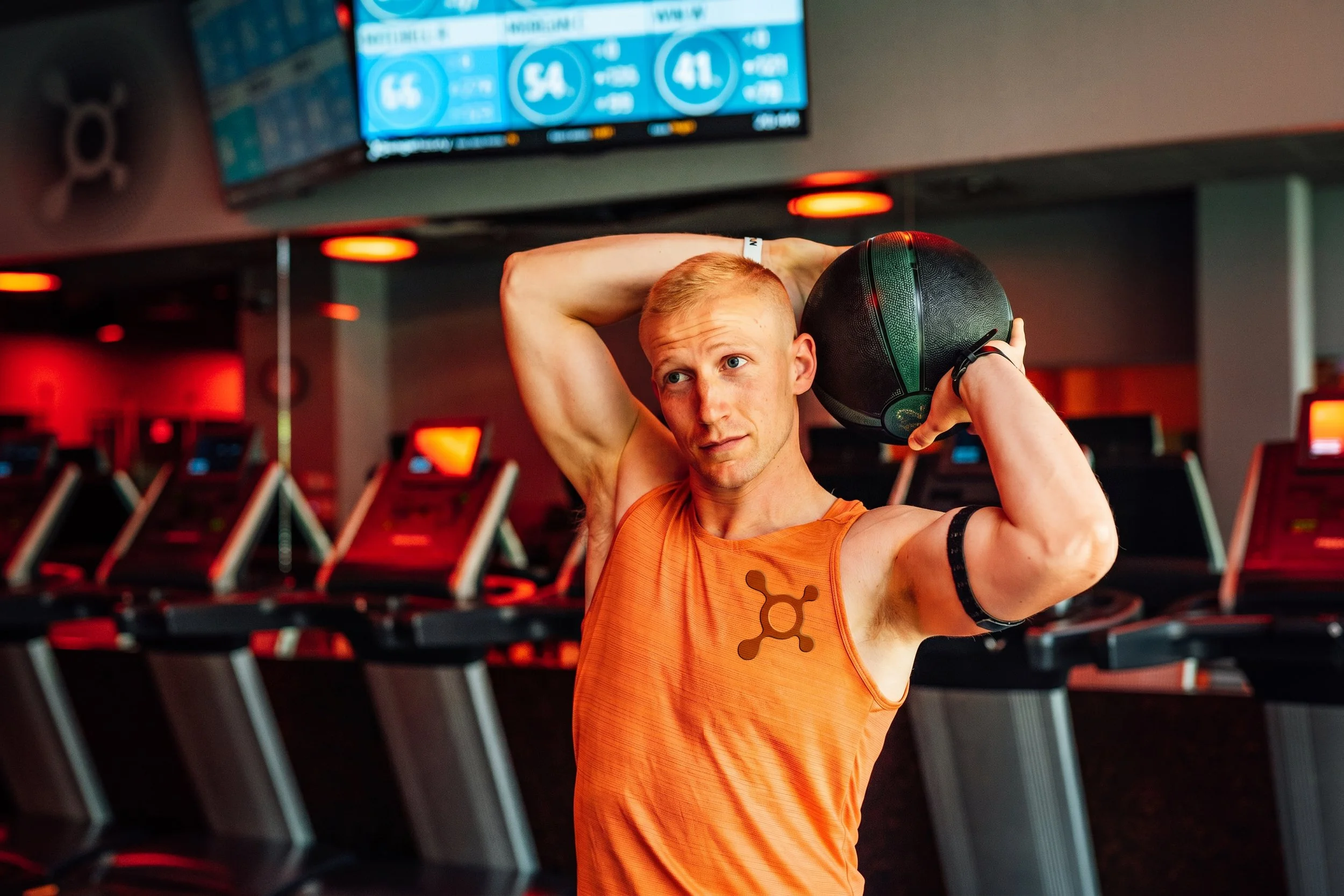 A man with blonde hair and blue eyes wearing an orange tank top holding a black medicine ball behind his head in a gym with red and black equipment.