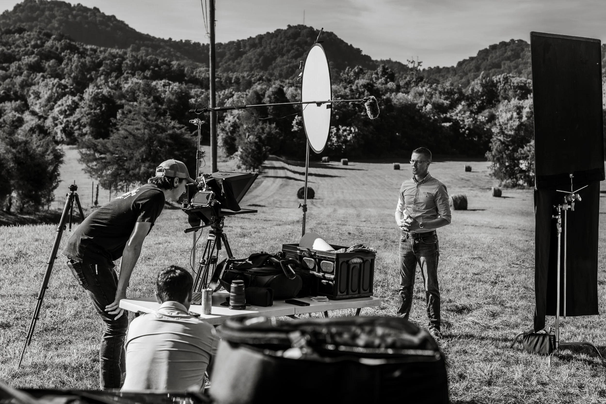 Black and white photo of an outdoor film or video shoot setup in a rural field with hay bales, surrounded by hills and trees. A man stands in front of a large panel, speaking or giving instructions. Multiple crew members are nearby, some operating equipment such as a camera and a microphone, with various gear and equipment on a table in the foreground.